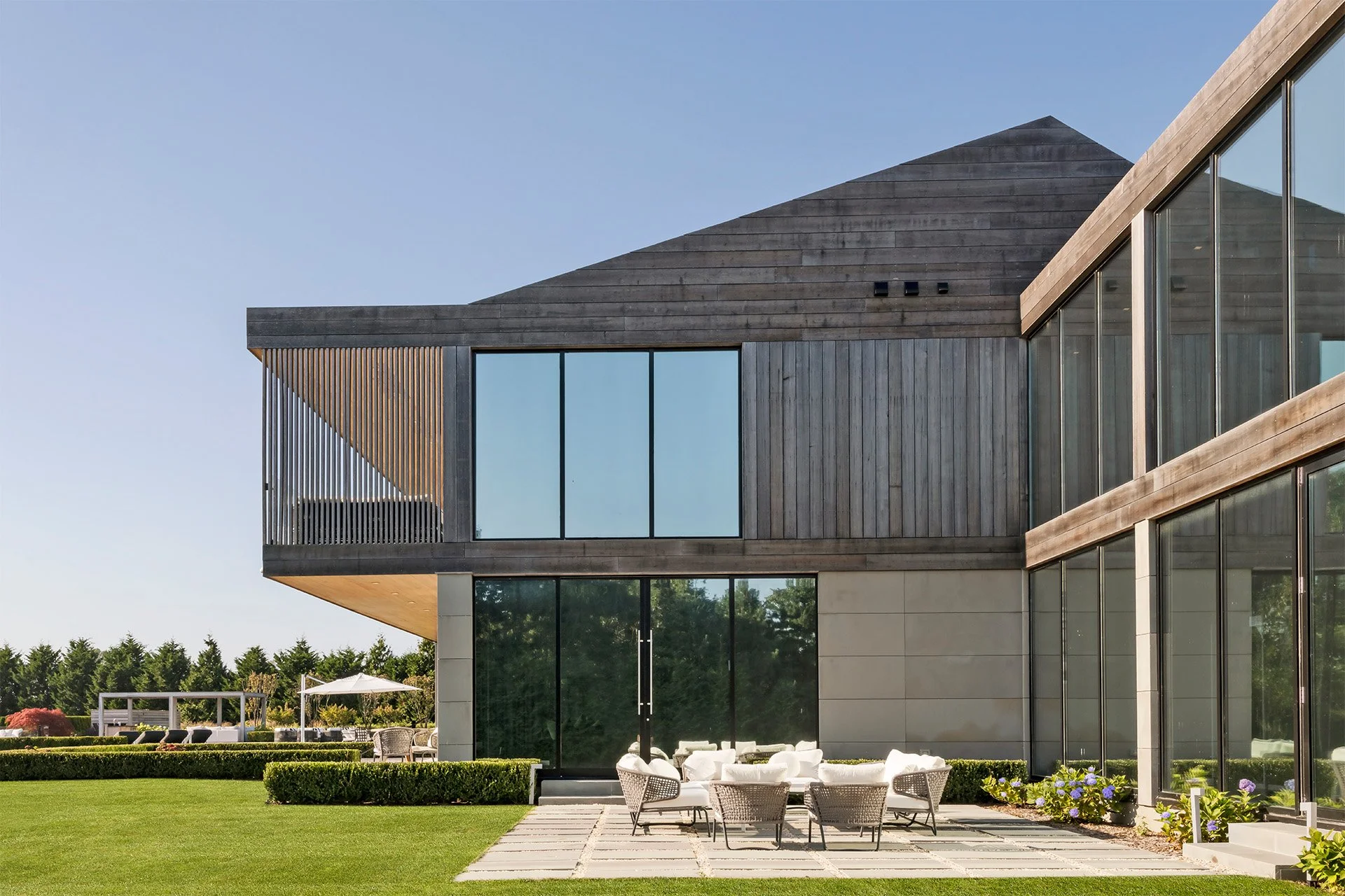 Modern house with large glass windows, wooden exterior, and a patio with white outdoor furniture, surrounded by a well-manicured lawn and greenery.