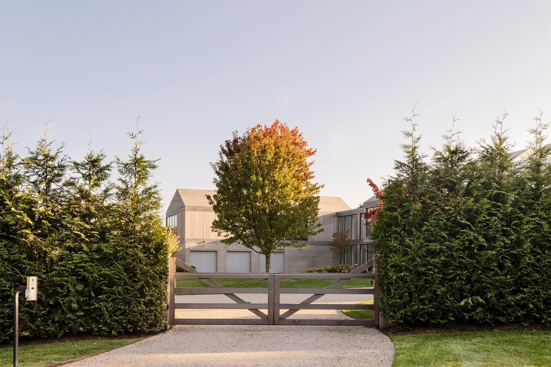 Modern residential house with a gray stone exterior, three garage doors, and large windows. A tree with autumn leaves stands in front, framed by tall green hedges on each side. There is a gravel driveway and a wooden gate in the foreground.
