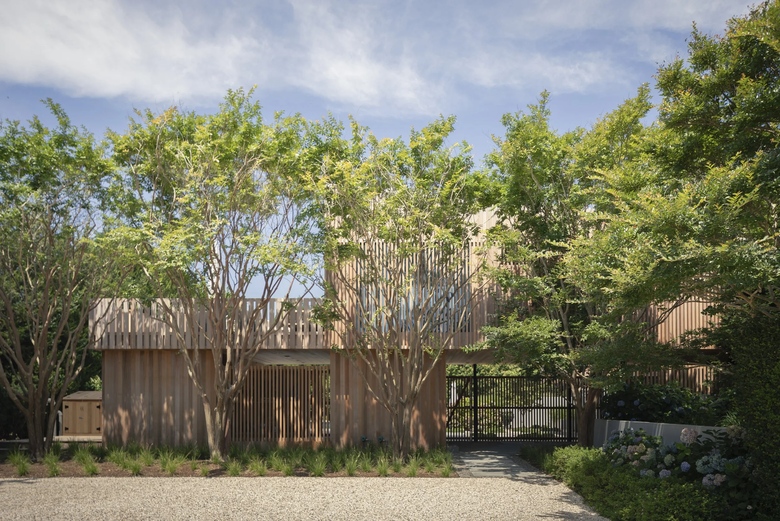 Modern house with wooden exterior, surrounded by trees and plants, with a gravel driveway and a black metal gate.