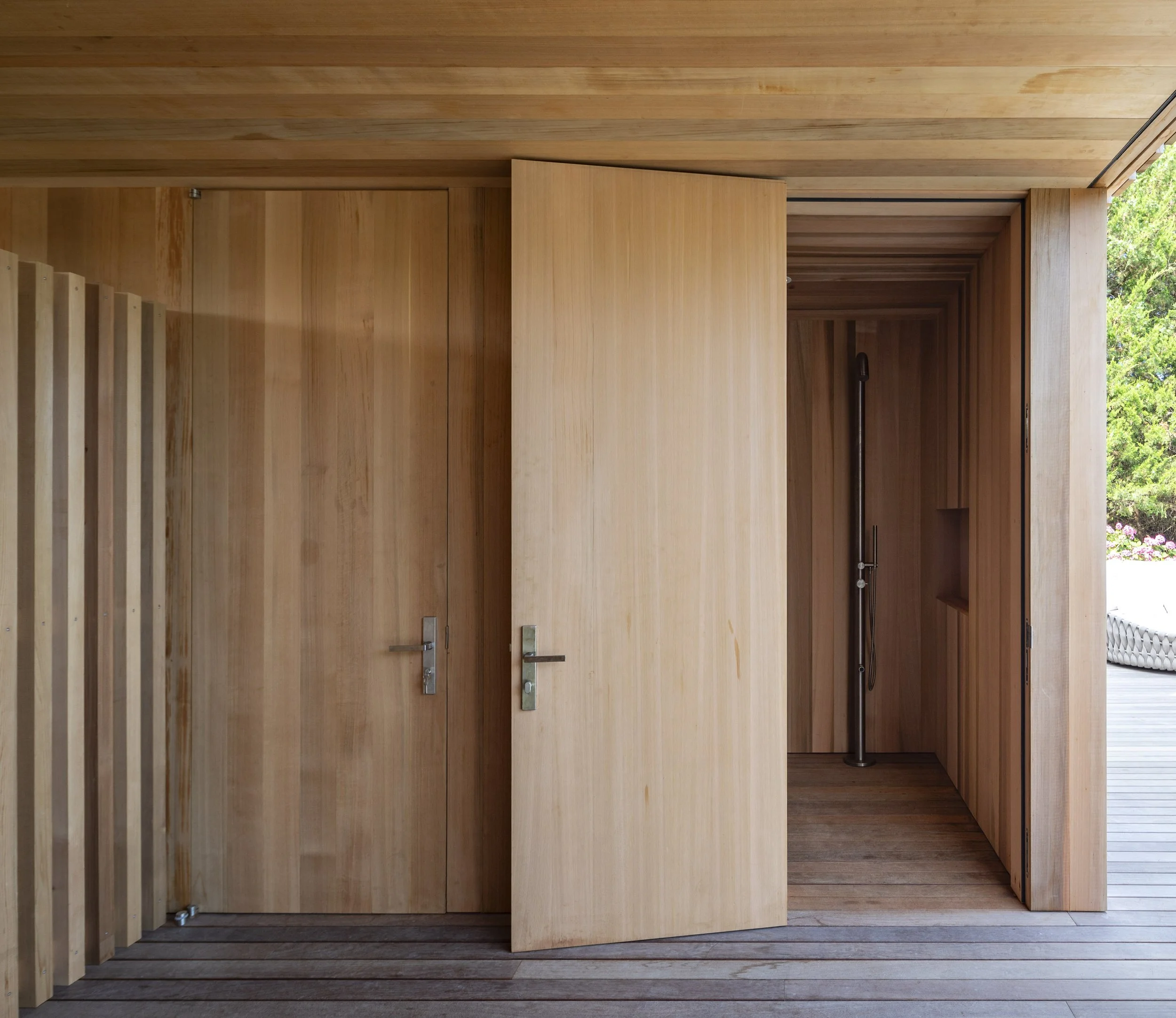 Interior view of a wooden shower with two doors, one open revealing a showerhead and controls on the wall.