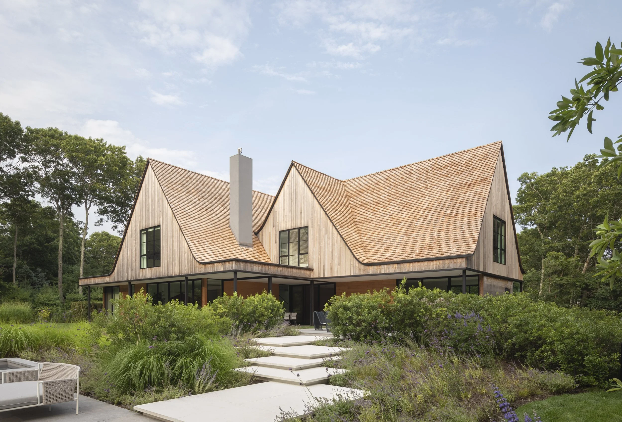 Modern house with a steep wooden shingle roof, large windows, and surrounded by lush greenery and a landscaped garden.
