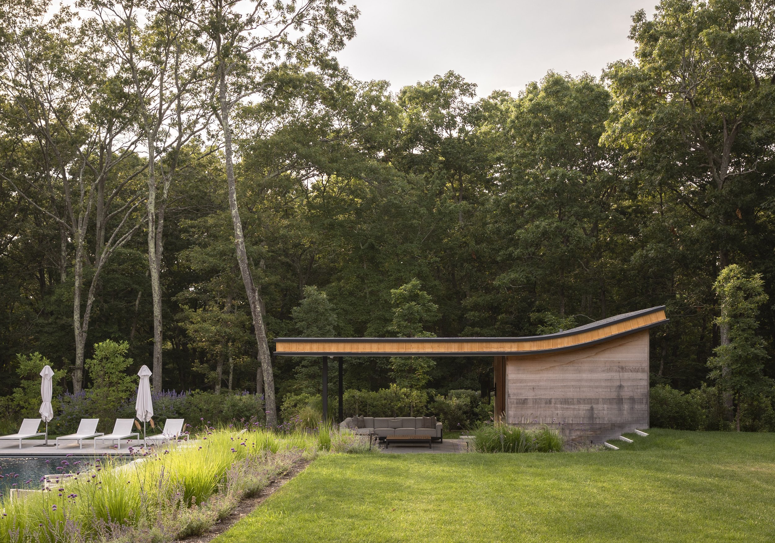 A modern backyard with a pool, white lounge chairs and umbrellas, a lush green lawn, a wooden outdoor shelter with outdoor furniture, surrounded by tall trees and greenery.