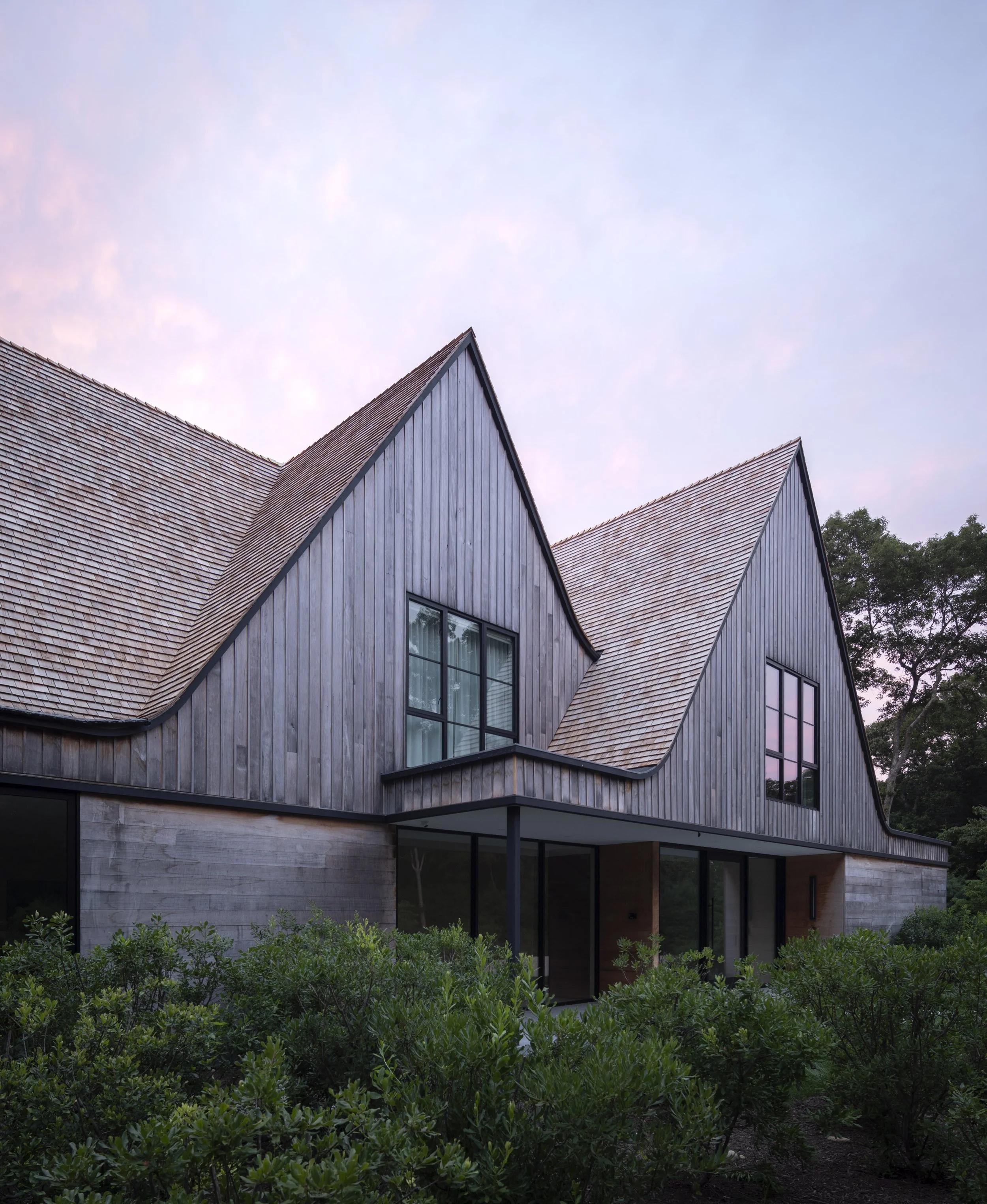 A modern house with a steep, curved wooden roof, large windows, surrounded by greenery at dusk.