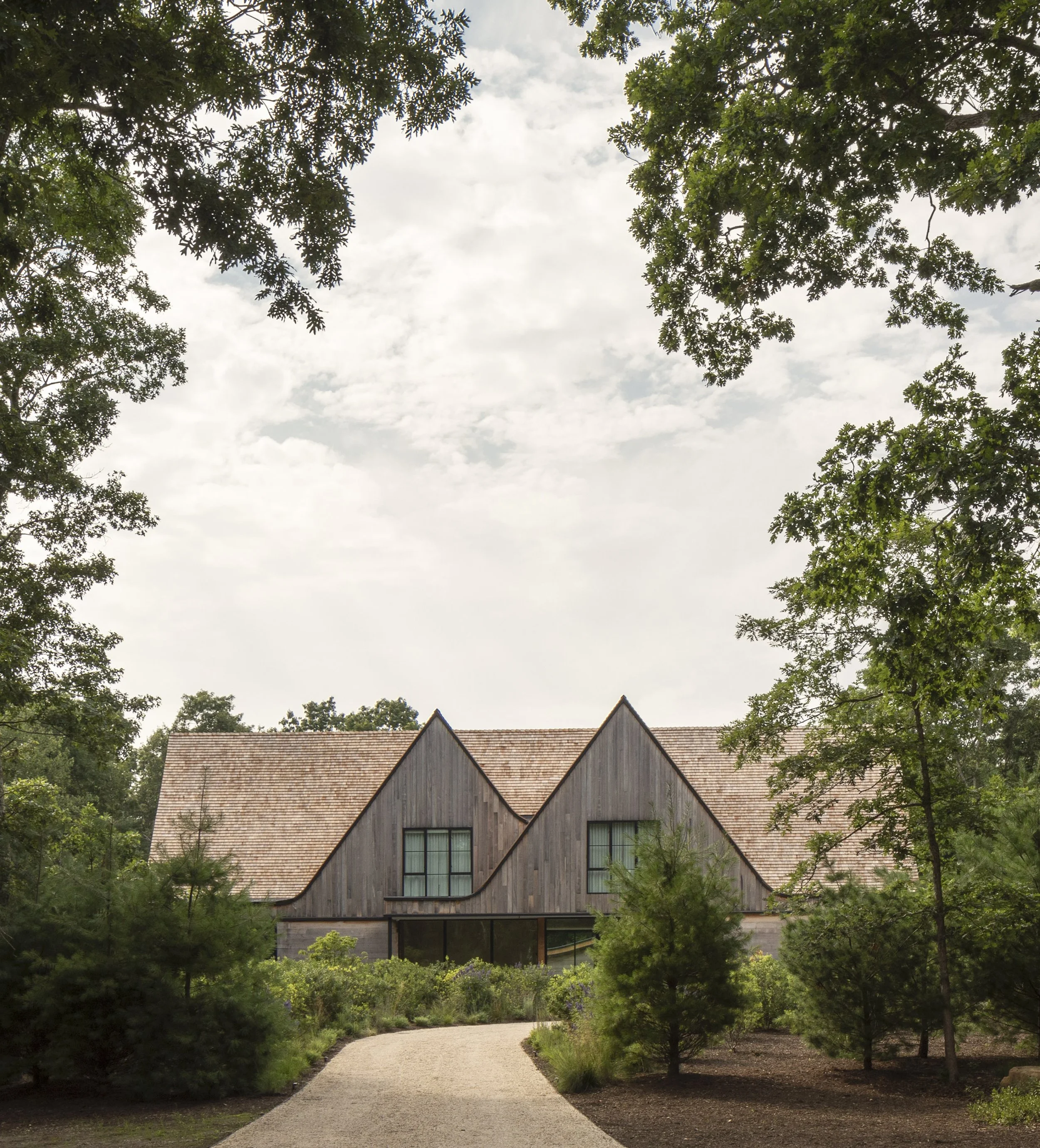 A modern house with wooden exterior and steep gable roofs, surrounded by trees and greenery, with a gravel pathway leading to it.