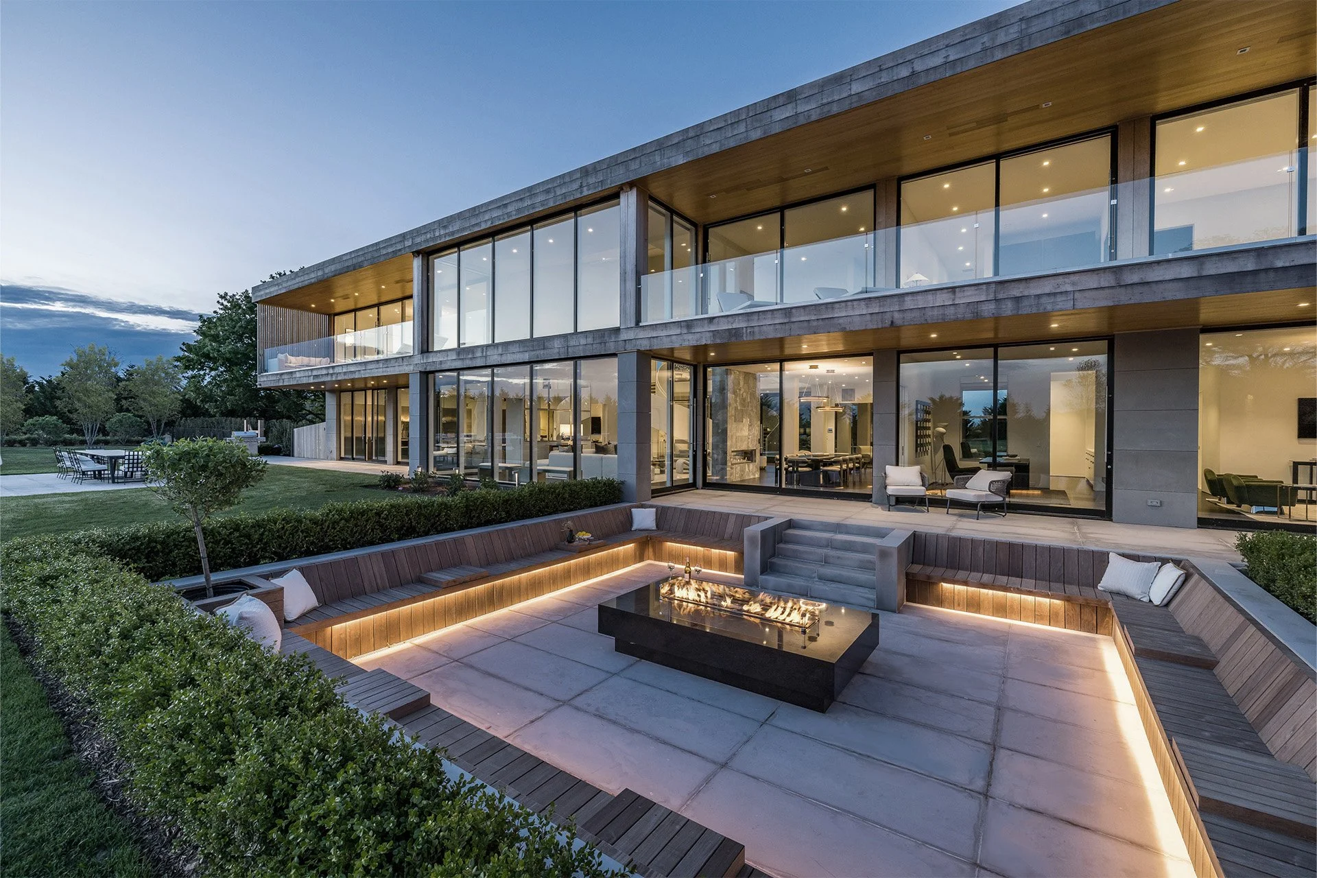 Modern multi-story house with large glass windows and outdoor patio area featuring built-in seating and a fire pit at dusk.