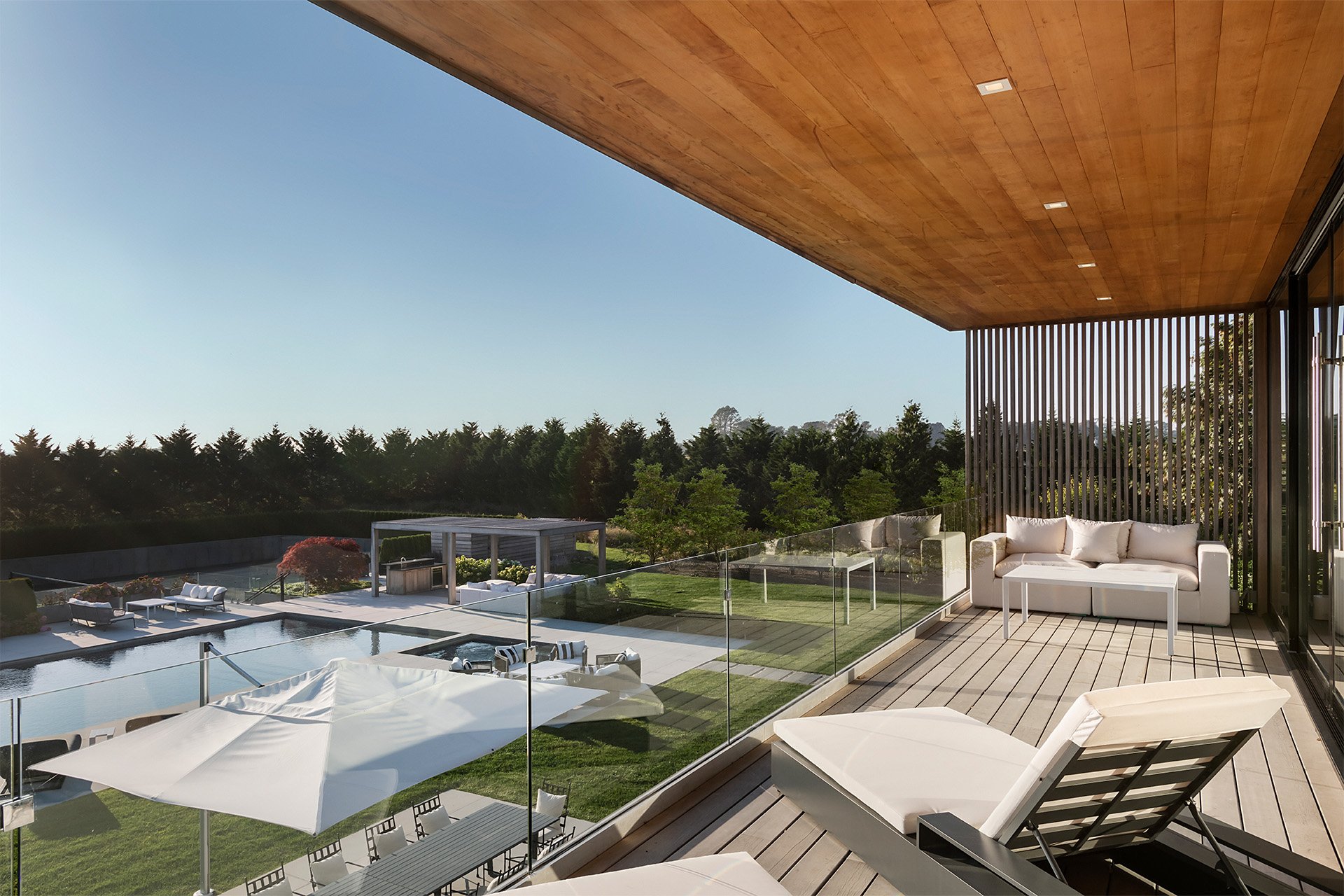 Modern balcony with white sofa, lounge chair, glass railing, outdoor pool area, trees, and clear blue sky.