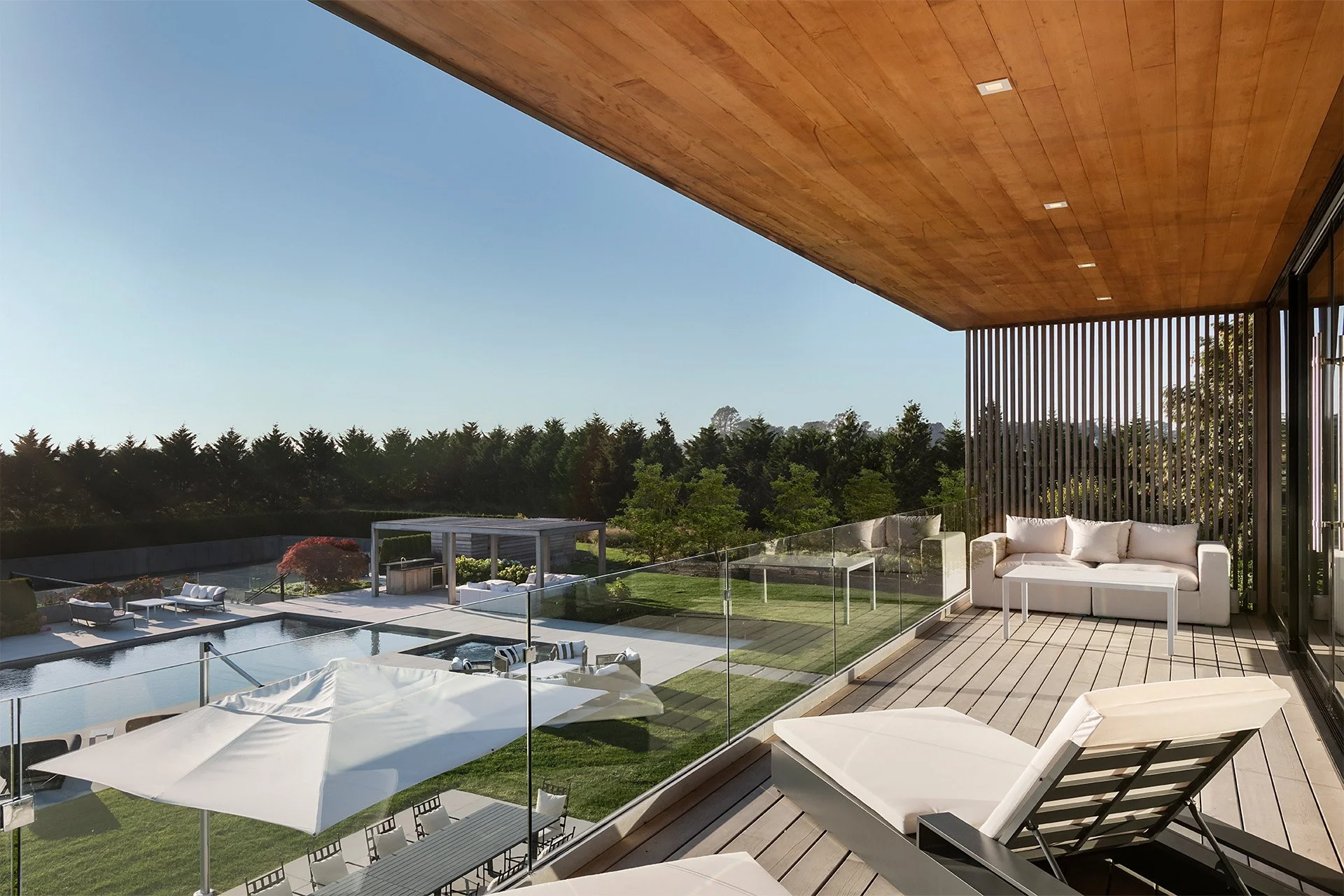 Luxury balcony with white outdoor furniture, glass railing, swimming pool, landscaped yard, and trees in the background under a clear sky.