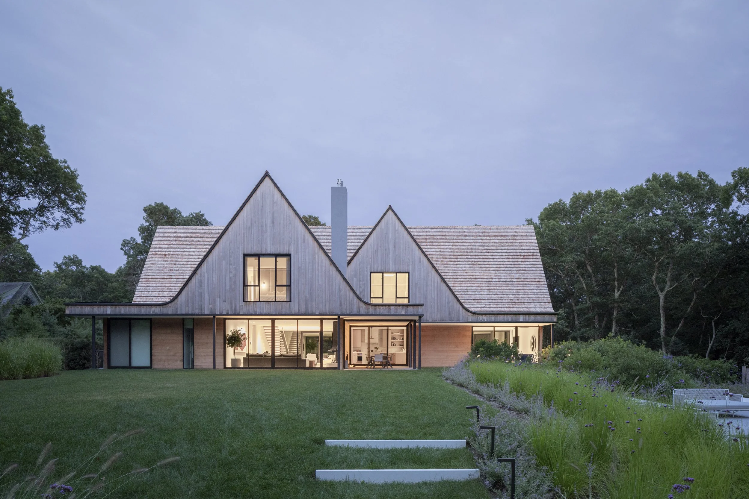 Modern house with a wooden exterior and large glass windows, situated in a lush green yard with trees in the background, during twilight.