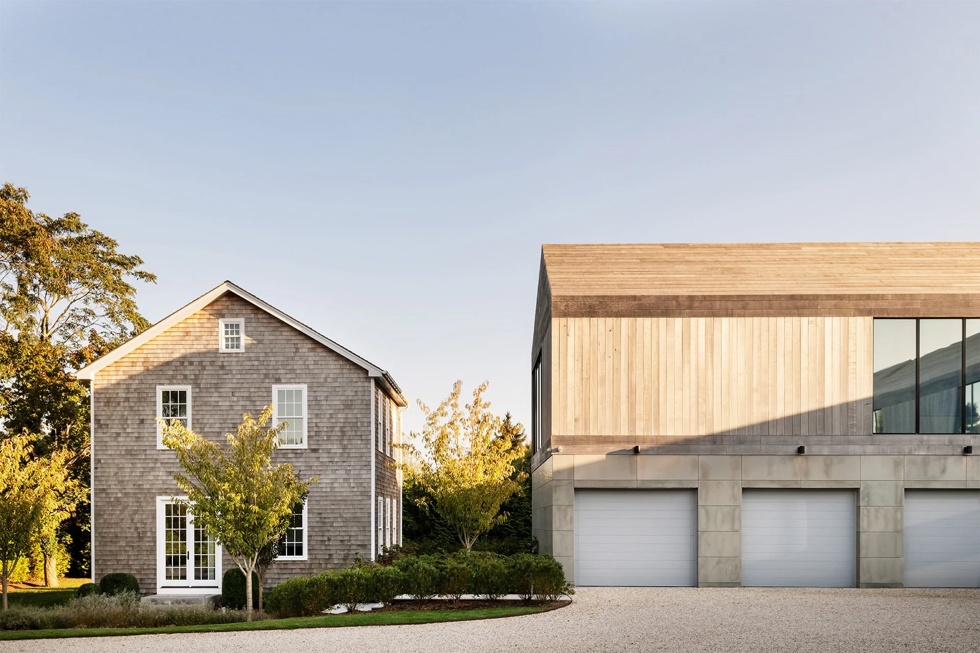 Two modern houses side by side, one with traditional wooden shingle exterior and the other with a contemporary design featuring wood and concrete, surrounded by green trees and a gravel driveway.