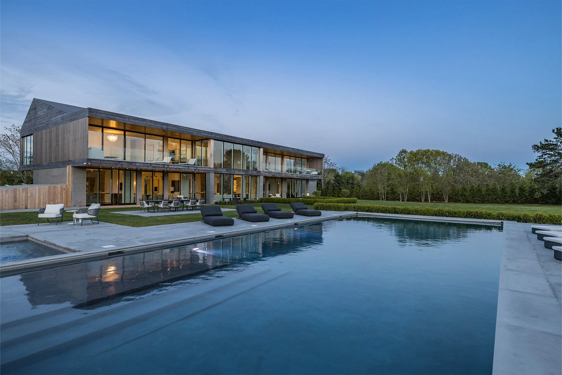 A modern two-story house with large glass windows and a swimming pool in the backyard during dusk, with outdoor seating and greenery in the background.