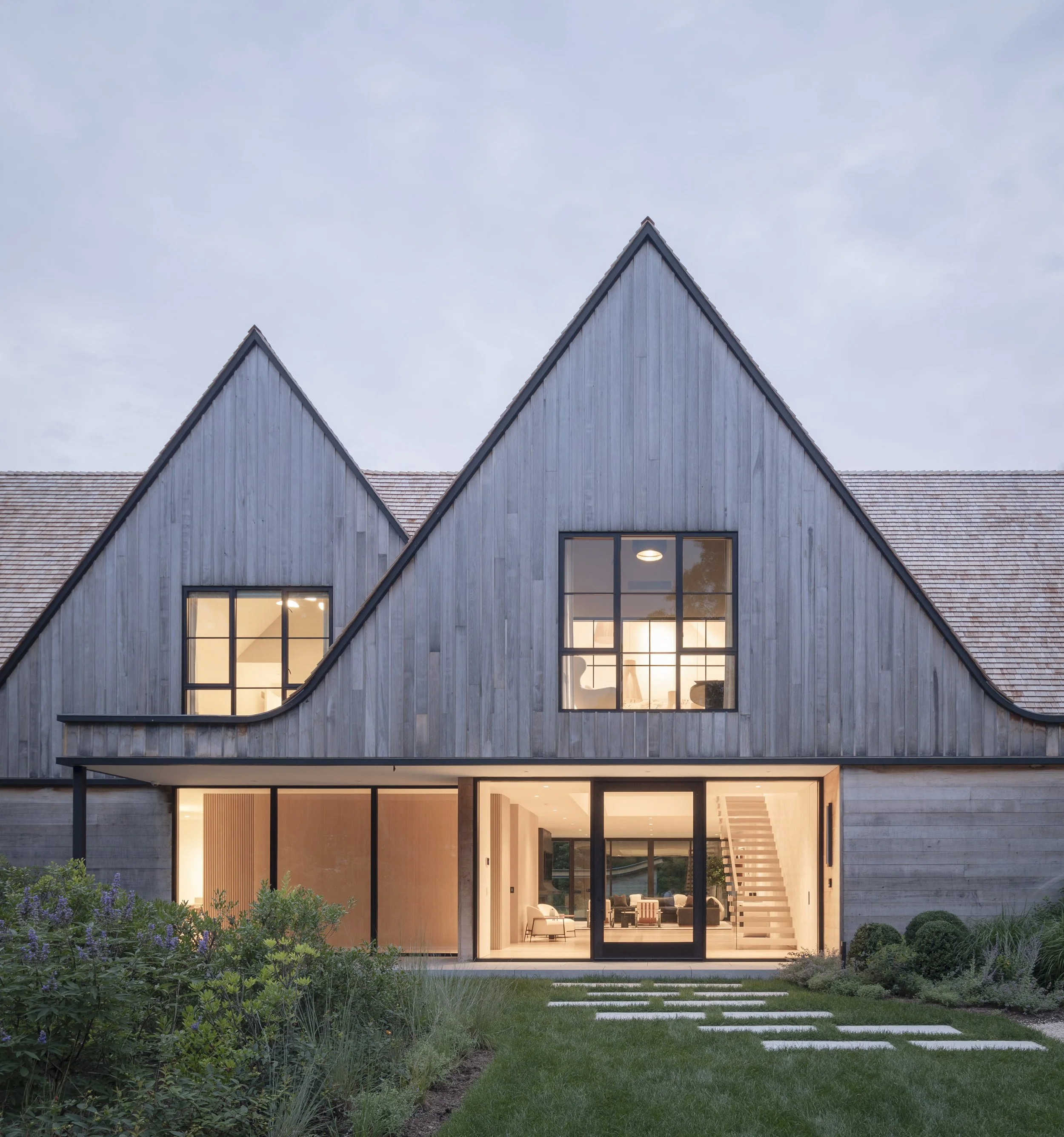 Modern house with gray wooden exterior, large glass doors and windows, surrounded by landscaped garden.