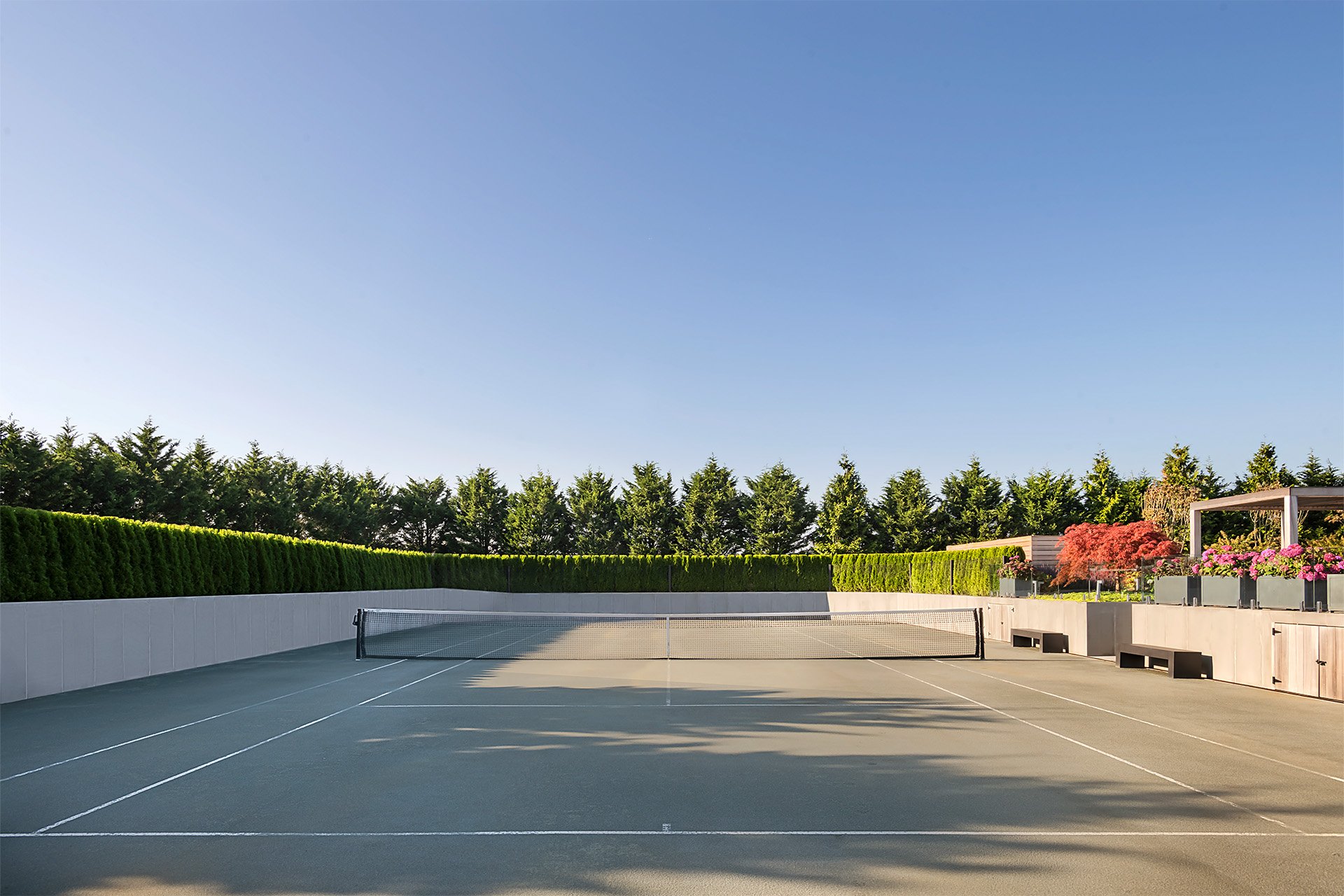 Empty outdoor tennis court with a net, surrounded by green hedges and trees, and a clear blue sky.