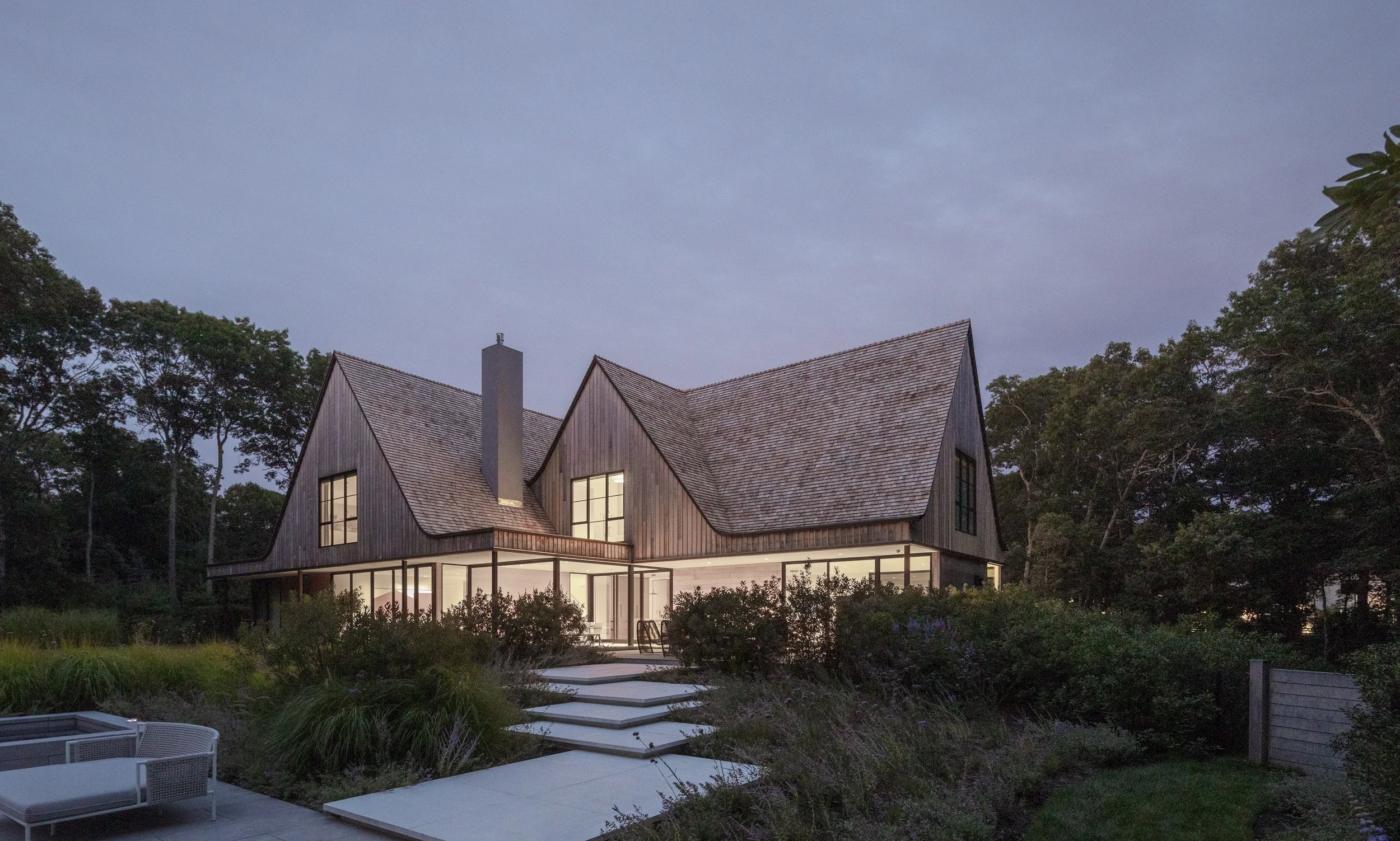 Modern house with large windows and wooden exterior, surrounded by lush greenery, at dusk.