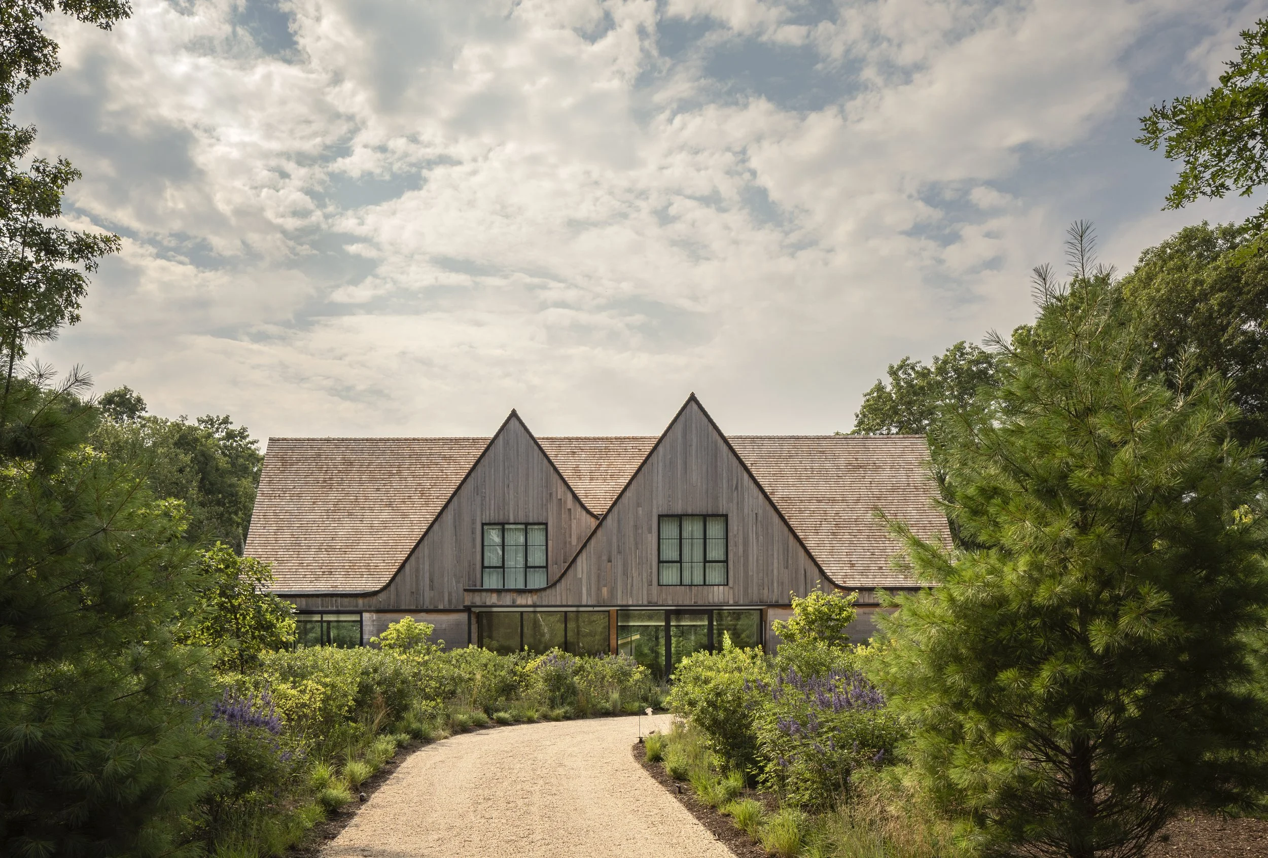 A rustic house with a steep wooden roof and large windows, surrounded by lush greenery and a gravel pathway leading to the entrance.