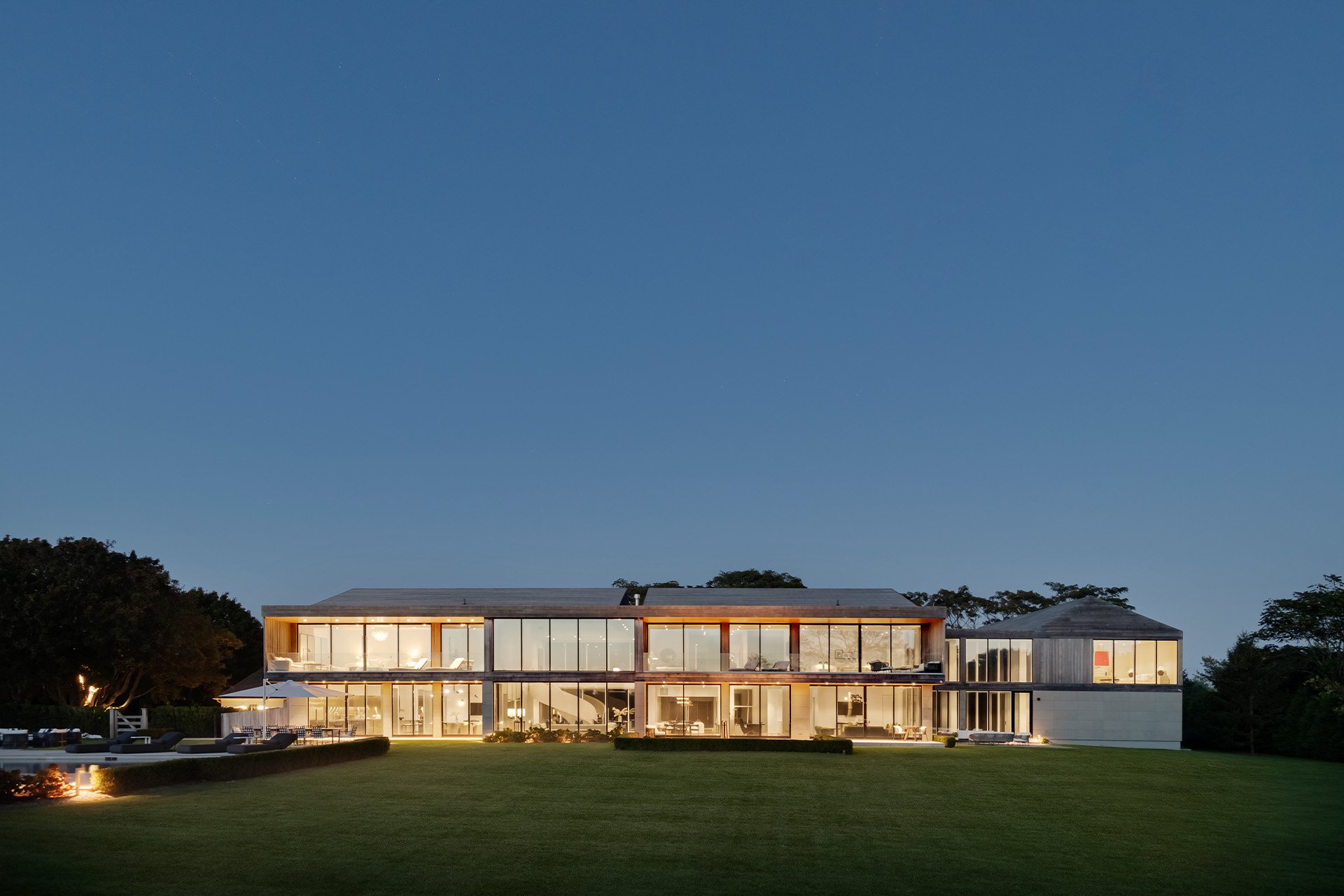 Modern two-story house with large glass windows and a well-manicured lawn, illuminated at night.
