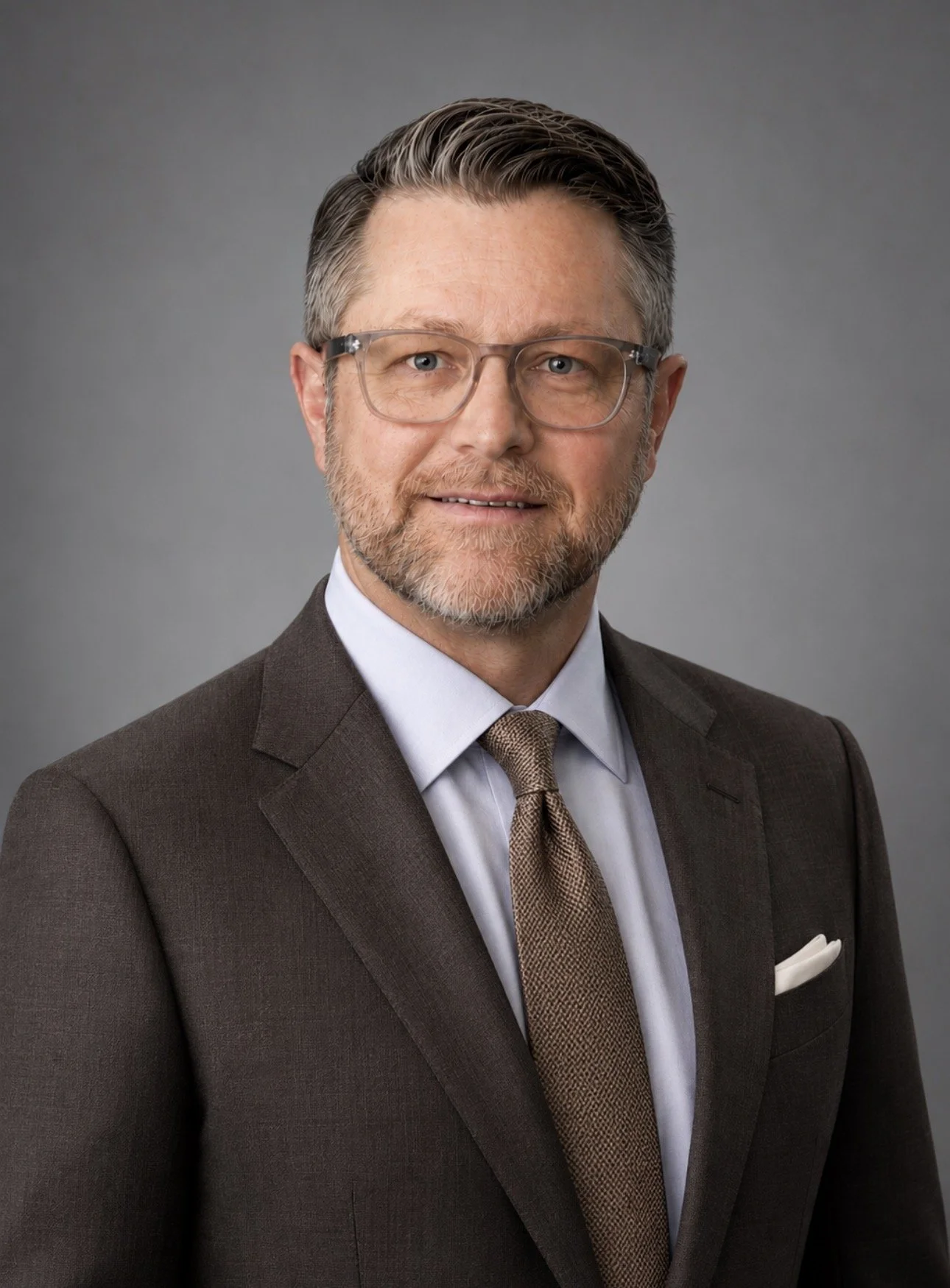 A middle-aged man with glasses, a beard, and combed-back hair, wearing a blue collared shirt and a dark blazer, smiling against a neutral grey background.