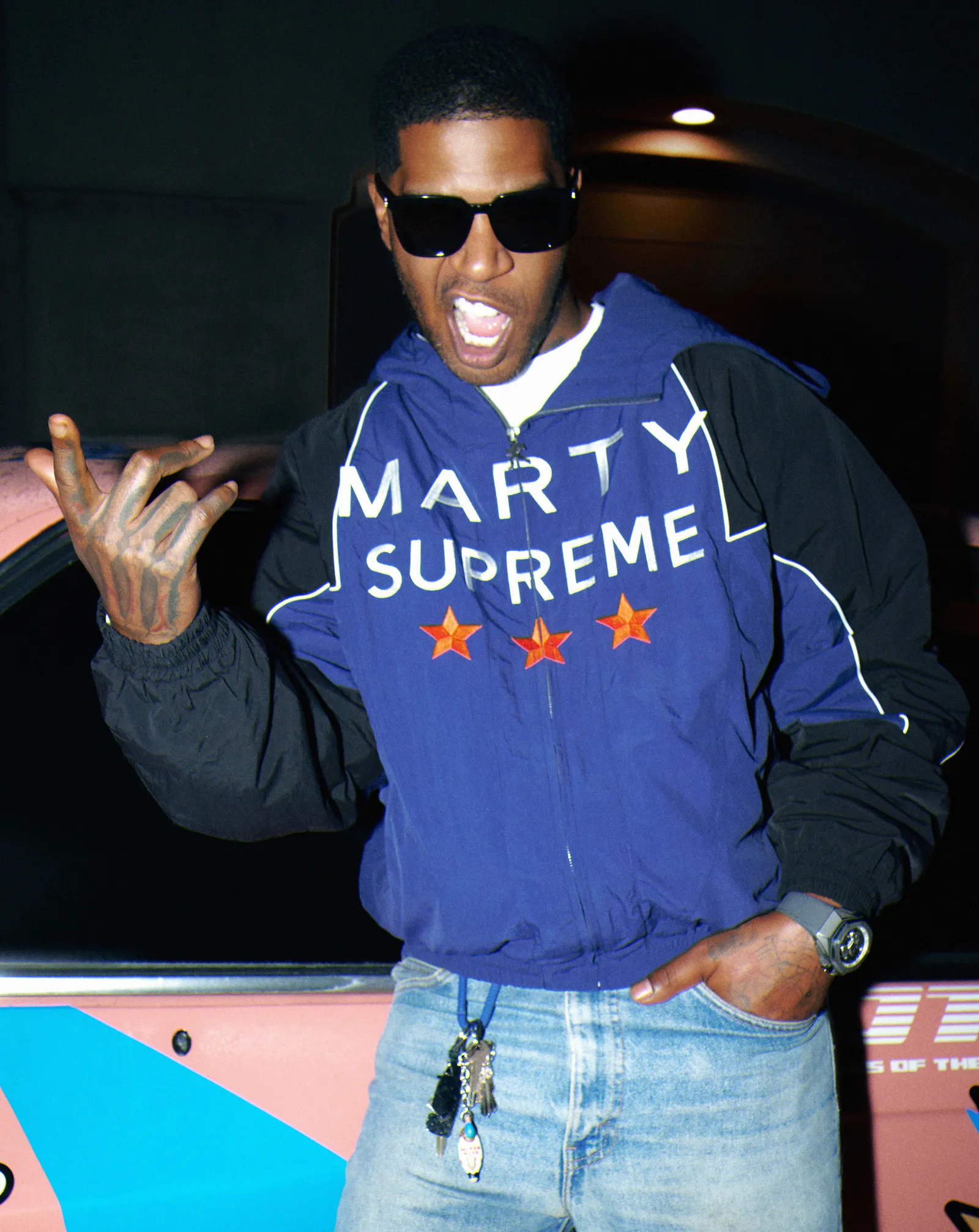 Black male adult wearing sunglasses and a blue Marty Supreme jacket with star graphics, posing at night beside a car with mouth open and hand raised.