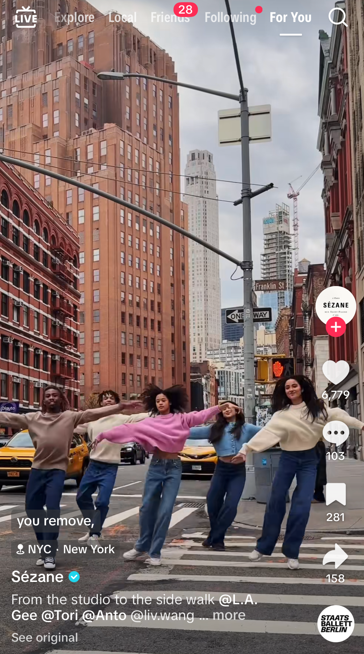 Five dancers move in unison across a New York City crosswalk, wearing casual sweaters and jeans with tall buildings and yellow taxis behind them.