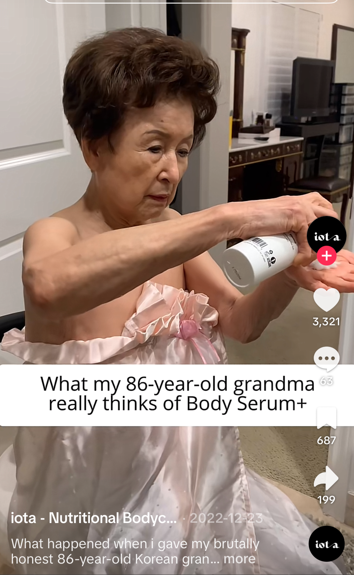 Elderly East Asian woman in a pink satin strapless dress pours white body serum lotion from a bottle into her palm while standing in a bedroom, with on-screen text reading “What my 86-year-old grandma really thinks of Body Serum+”.