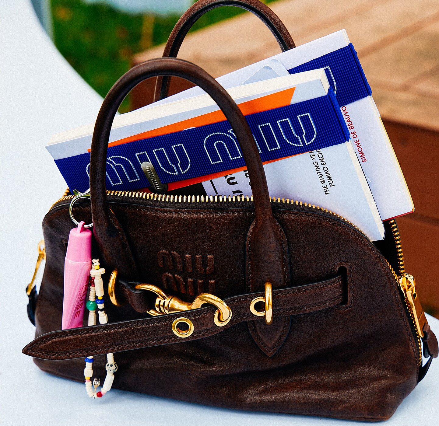 brown leather Miu Miu handbag with gold hardware, partially unzipped to reveal several books and magazines inside, including a blue “Miu Miu” branded band wrapped around them; the bag sits on a table with a pink lip gloss and beaded keychain