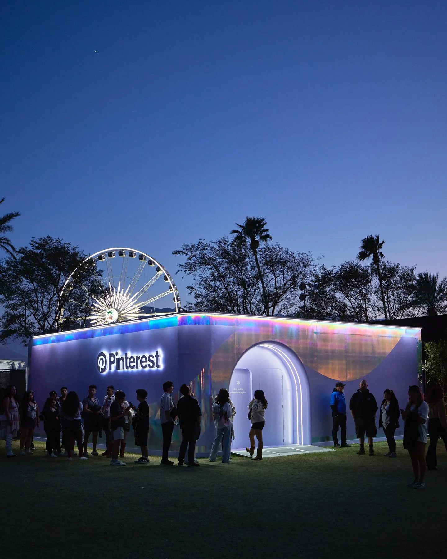 Outdoor evening scene at the Pinterest activation at Coachella, where a glowing iridescent pop-up structure with a lit “Pinterest” logo stands on grass, as a diverse group of adults line up and gather around the entrance.
