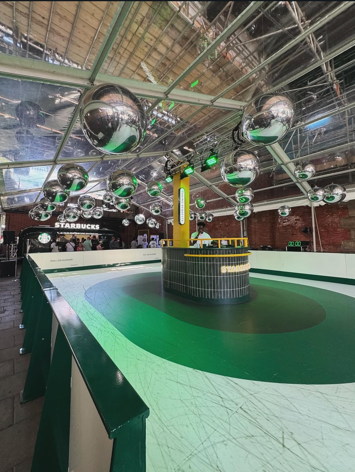 Indoor skating rink installation for Starbucks “Roll on Summer” event in Manchester, featuring a green circular rink, central DJ booth, reflective silver hanging spheres, and a Starbucks café bar with visitors in the background.