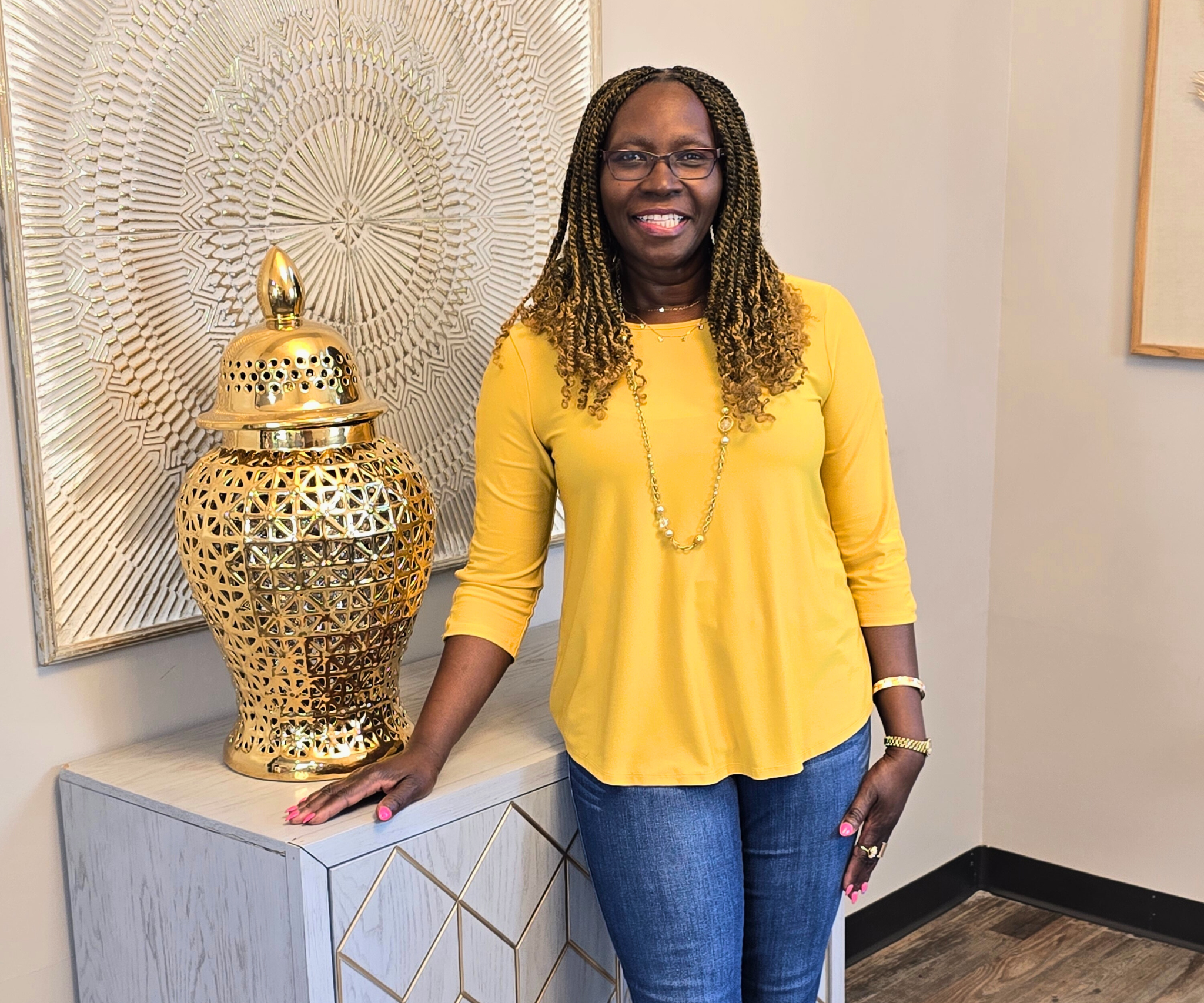 A woman dressed in a yellow top and blue jeans standing next to a decorative gold vase with a lattice design, in an interior space with wall art and decor.