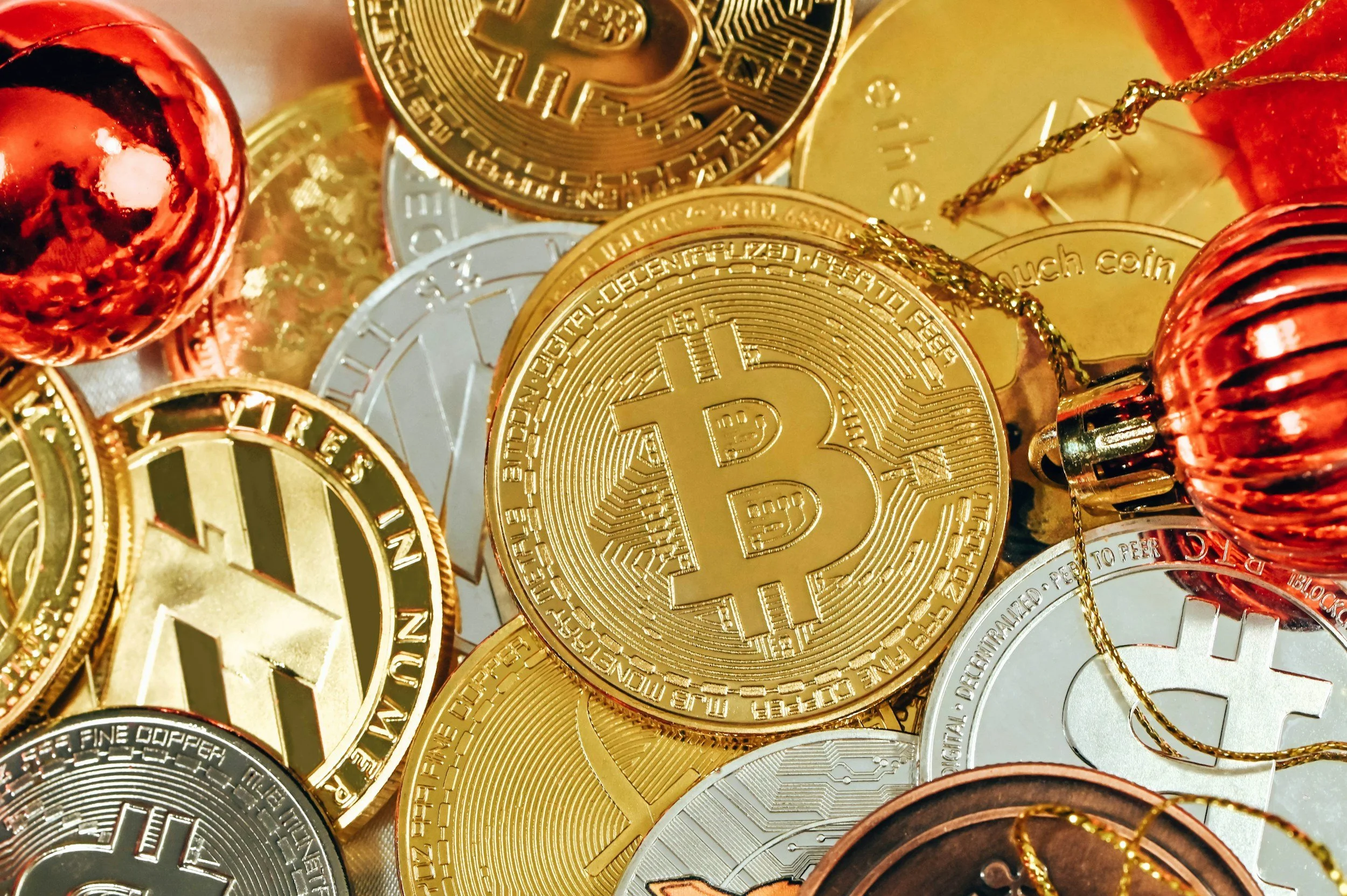 Close-up of various cryptocurrency coins, including Bitcoin, Litecoin, and Dogecoin, with some in gold, silver, and copper colors. Some coins are decorated with small Christmas ornaments.