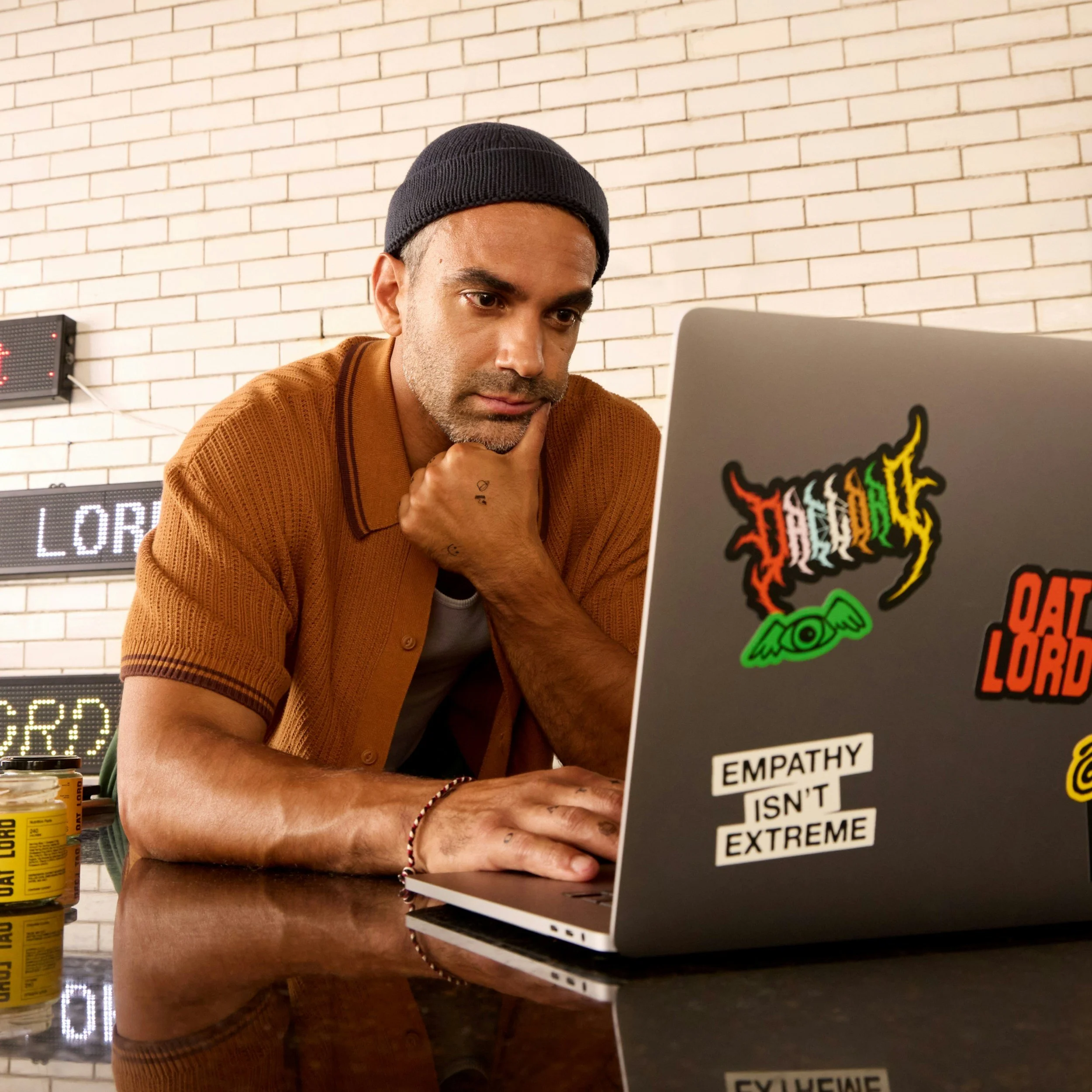 Man with gray beard wearing a black beanie and orange shirt working on a laptop with stickers, including one that says "Empathy Isn't Extreme," in a room with a brick wall and electronic sign in the background.