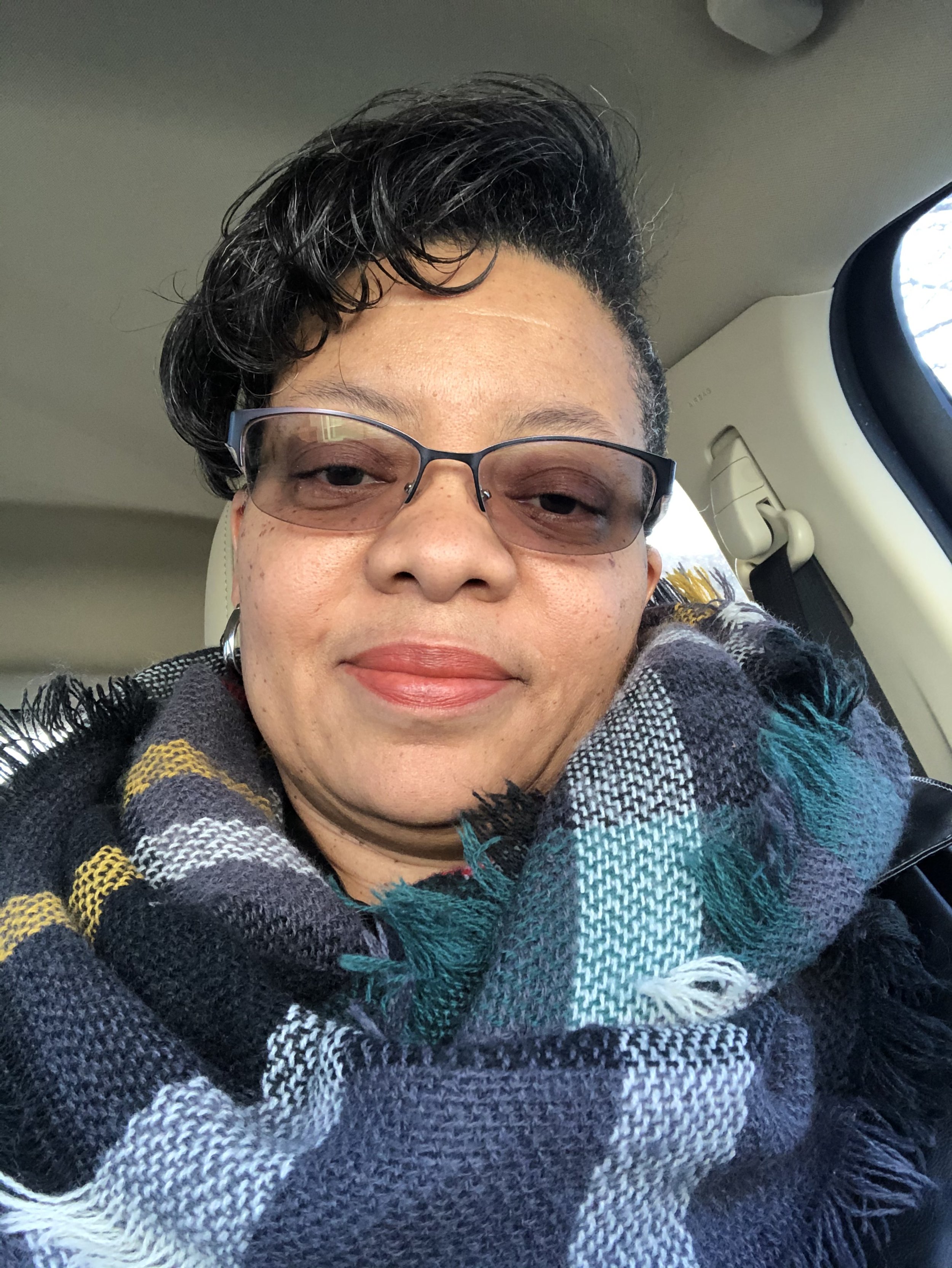 A woman taking a selfie inside a vehicle, wearing glasses, a plaid scarf, and earrings, with a slight smile.