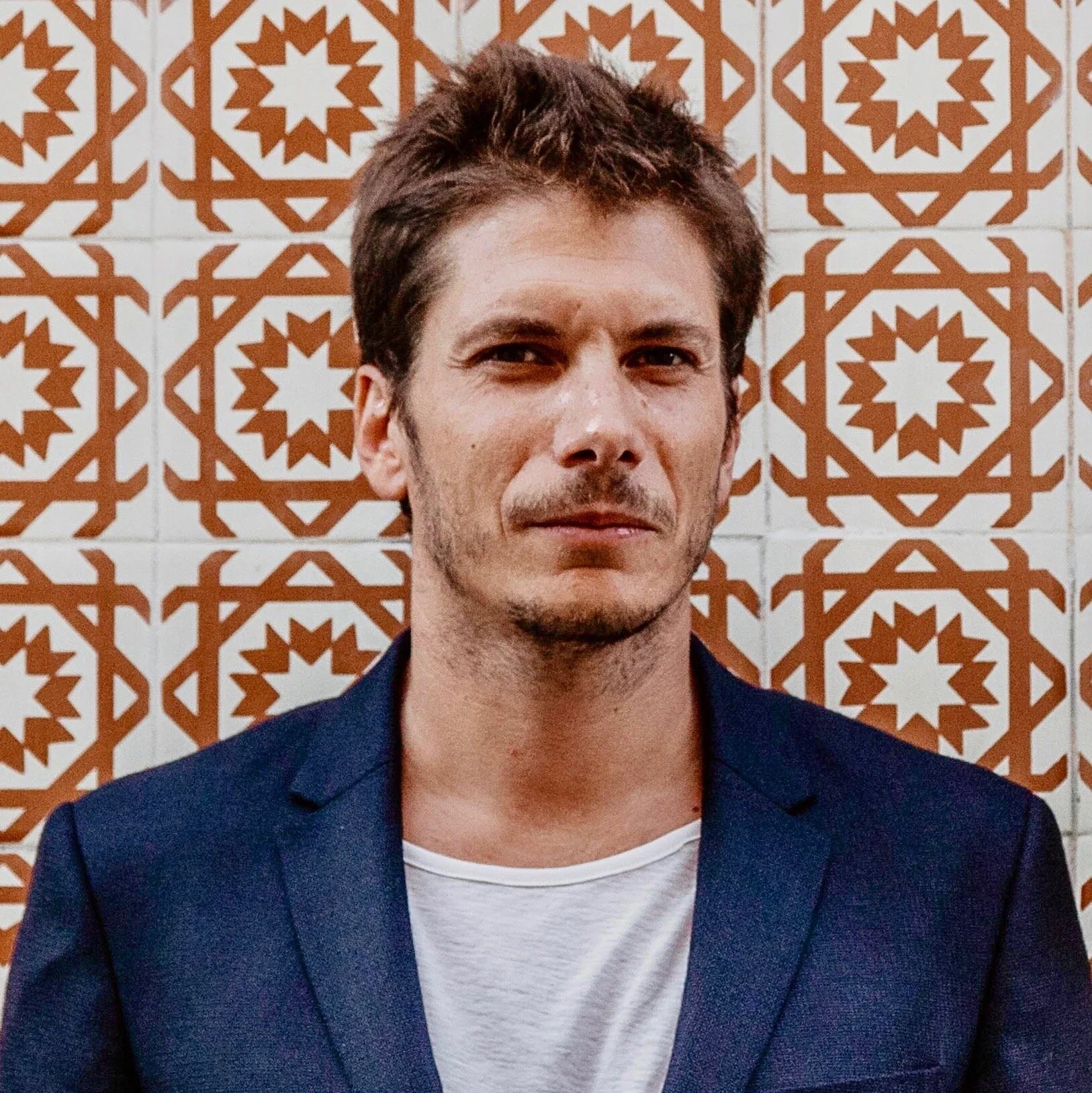 Homem com cabelo castanho e barba, vestindo terno azul e camiseta branca, posando em frente a azulejos dourados com padrão geométrico.