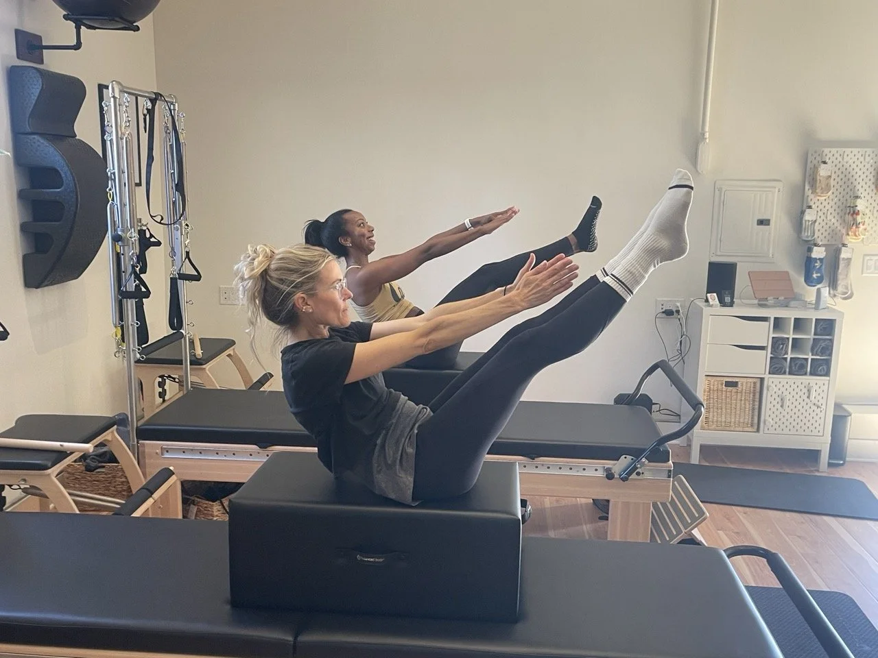 Two women exercising on Pilates reformer machines in a fitness studio, with one woman in the foreground and the other in the background, both raising their arms and legs during a workout.