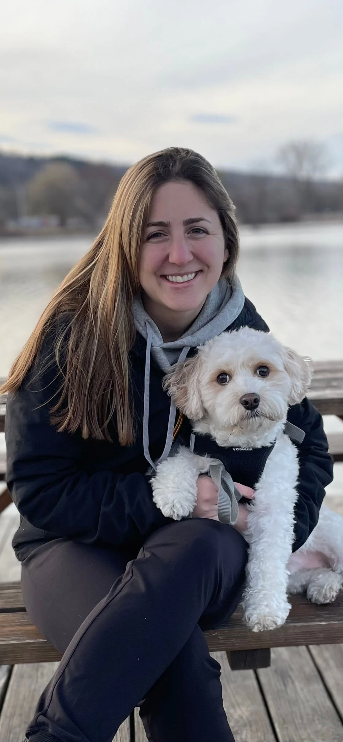Riya Burke, Math tutor, New York tutor, sitting on a wooden bench outdoors, holding a small white dog with curly fur in her lap, near a body of water with trees in the background.