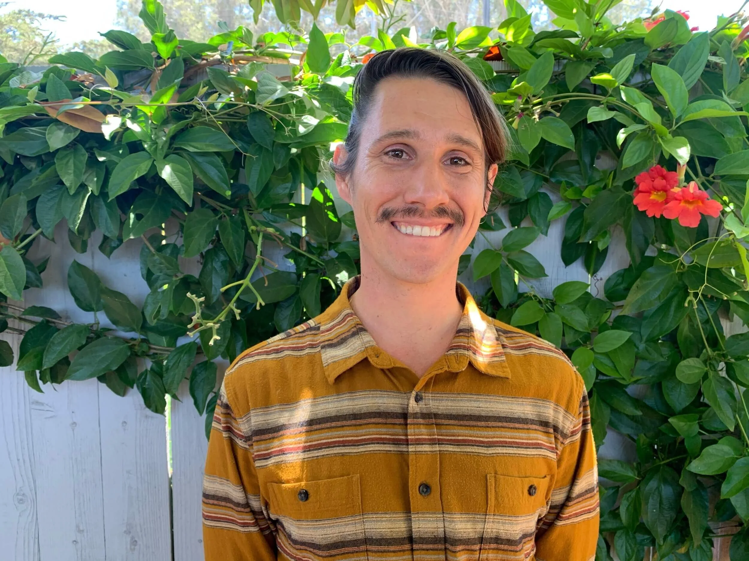 Jonathan Wilcox, Santa Barbara tutor, focusing on Mathematics tutoring, Physics tutoring, SAT tutoring & ACT tutoring, wearing a mustard yellow and brown striped shirt, standing outdoors in front of a lush green leafy plant with some red flowers.