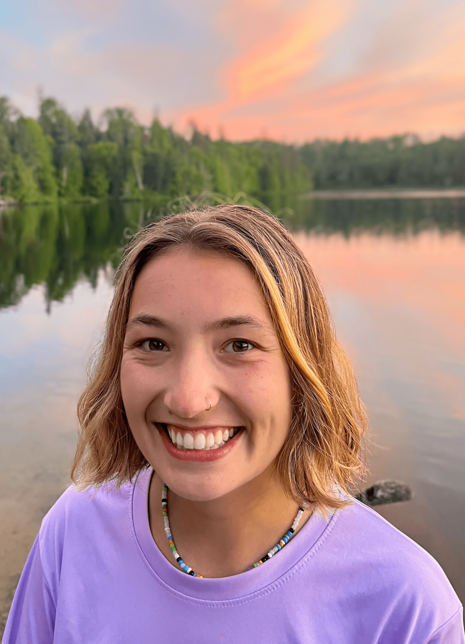 Emma Clark, Language Arts & Mathematics tutor, with shoulder-length blonde hair smiling at camera, standing near a lake during sunset with a pink and orange sky and trees reflected in the water.