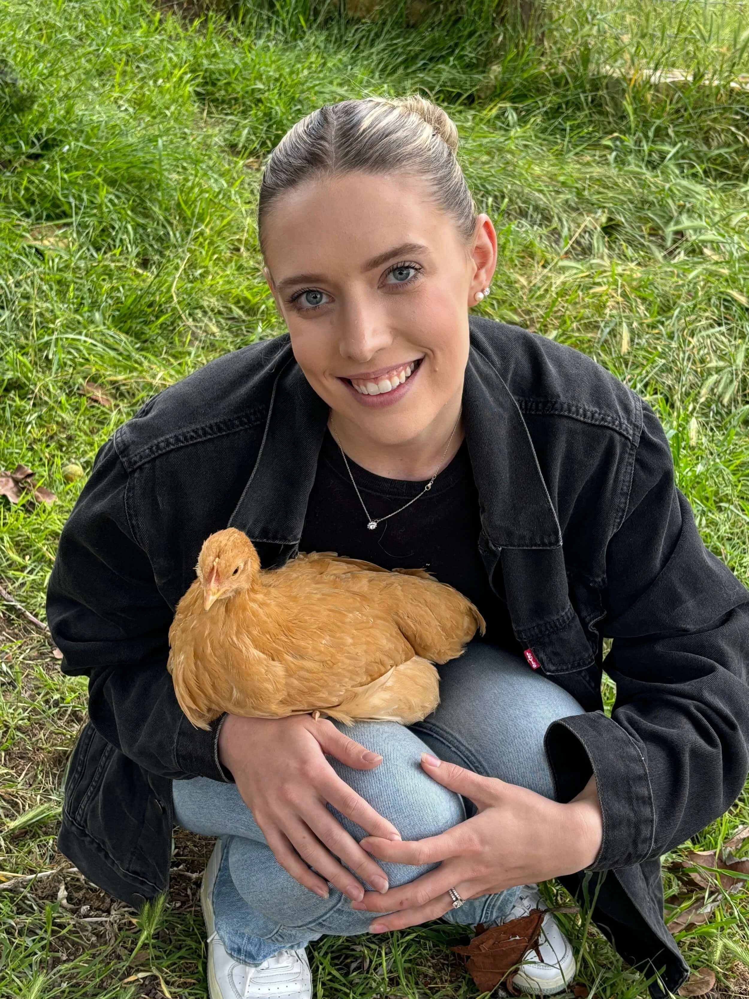 Dela Morgan, Biology and Chemistry tutor, in the grass, holding a light brown chicken on her lap, smiling at the camera. She is wearing a black jacket, light blue jeans, and white sneakers.