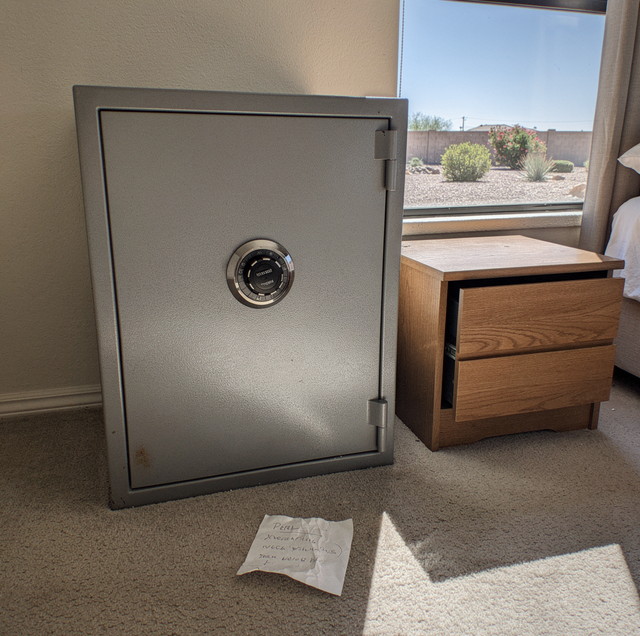 A gray safe with combination lock sits on carpet next to a wooden dresser near a window overlooking a desert landscape.