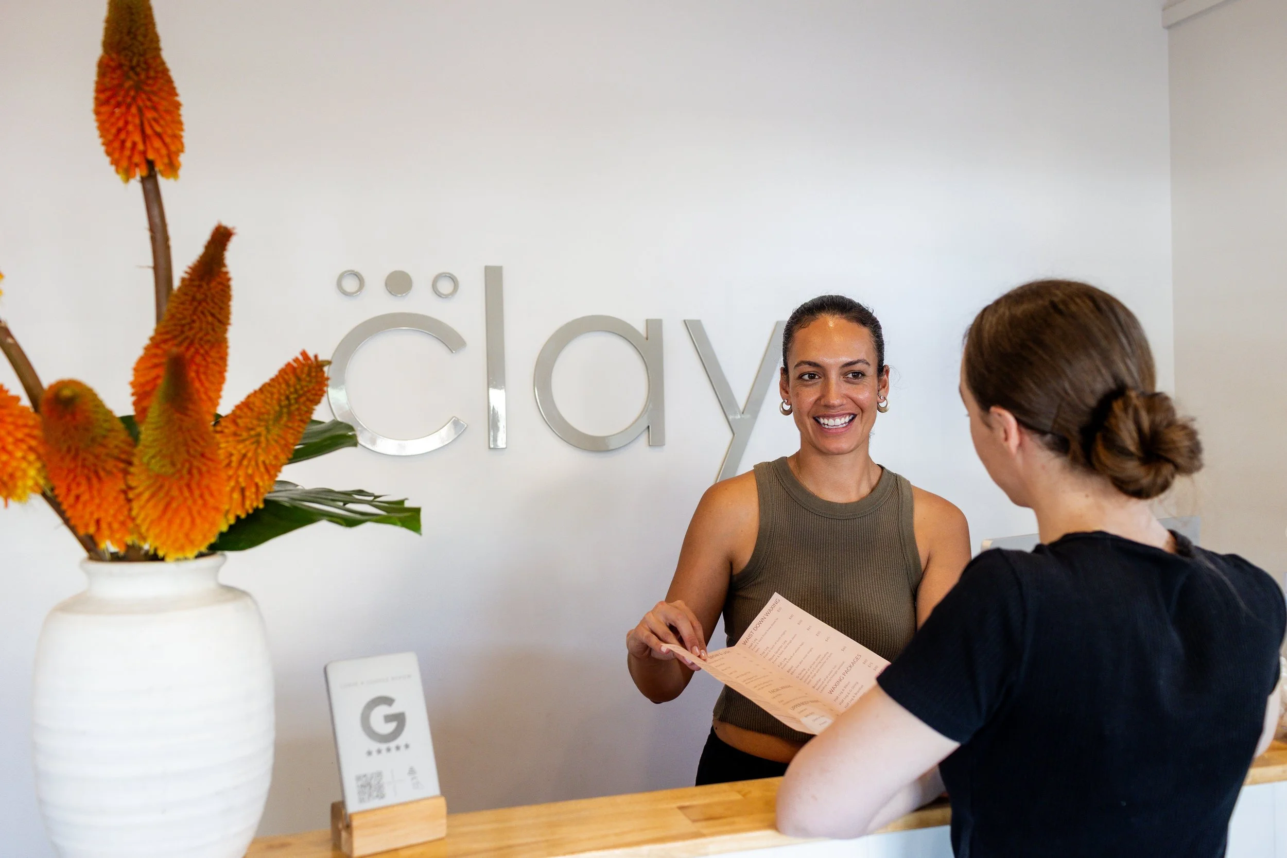 A woman with dark hair tied back, smiling and wearing a sleeveless green top, is handing a pink paper to a customer with brown hair tied in a low bun, wearing a black top, at a reception desk in a place called 'Cílay.' A large white vase with orange