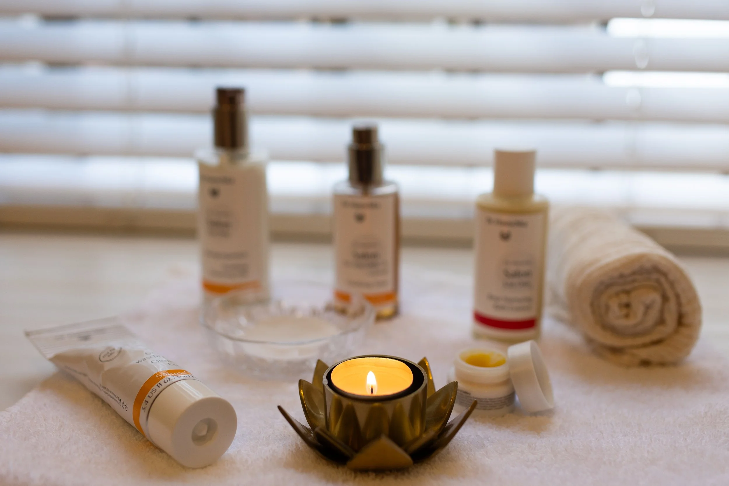 Spa or skincare set with three bottles, a rolled towel, a small jar, a glass bowl, a candle in a lotus-shaped holder, and a tube of cream on a white towel in front of window blinds.