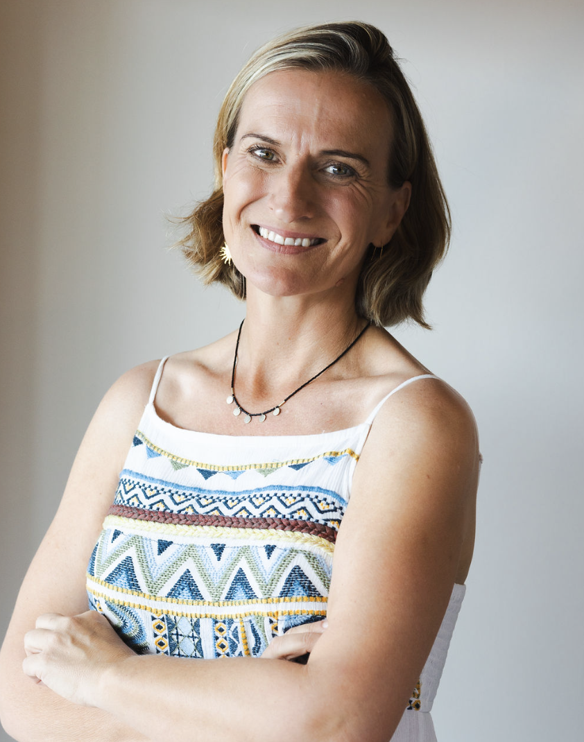 A woman with shoulder-length light brown hair, smiling with crossed arms, wearing a white sleeveless top with colorful embroidered patterns, a black necklace, and gold earrings, standing against a plain light-colored background.