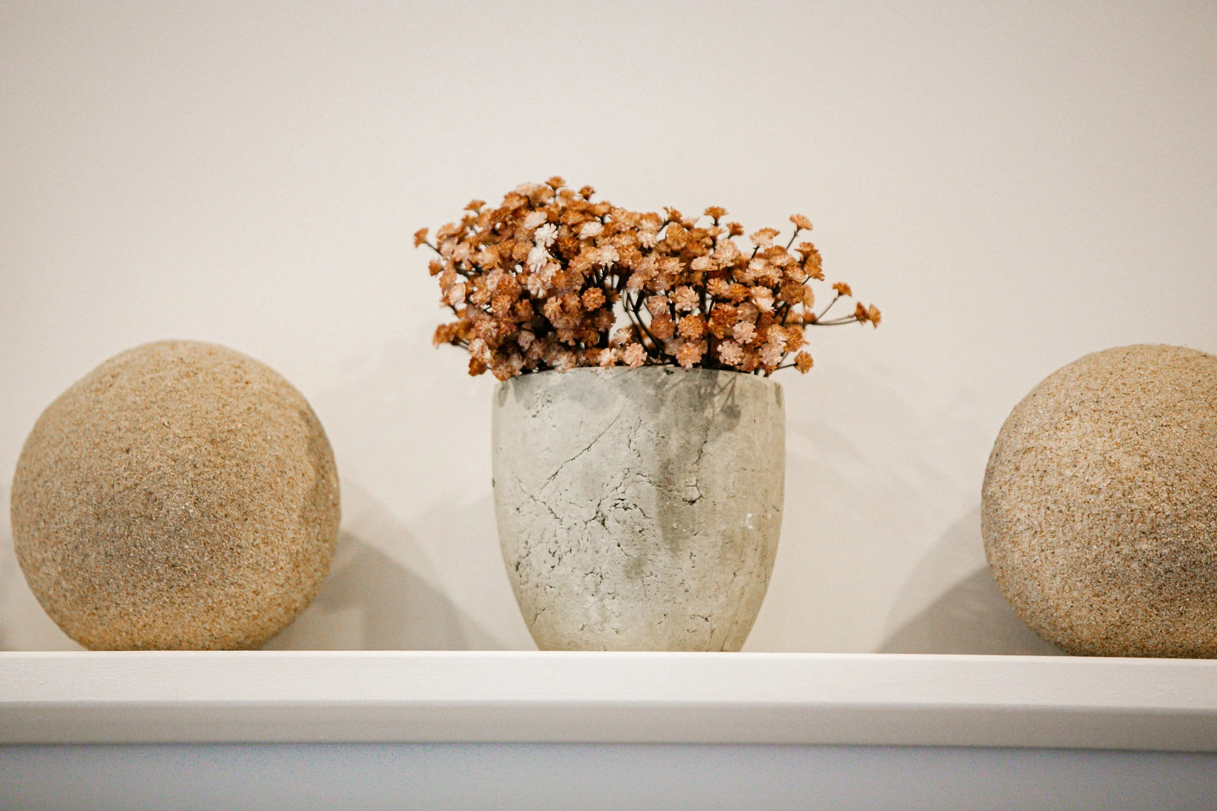 Decorative arrangement of two rounded stones flanking a concrete vase with dried pink flowers, all on a white shelf, against a plain light-colored wall.