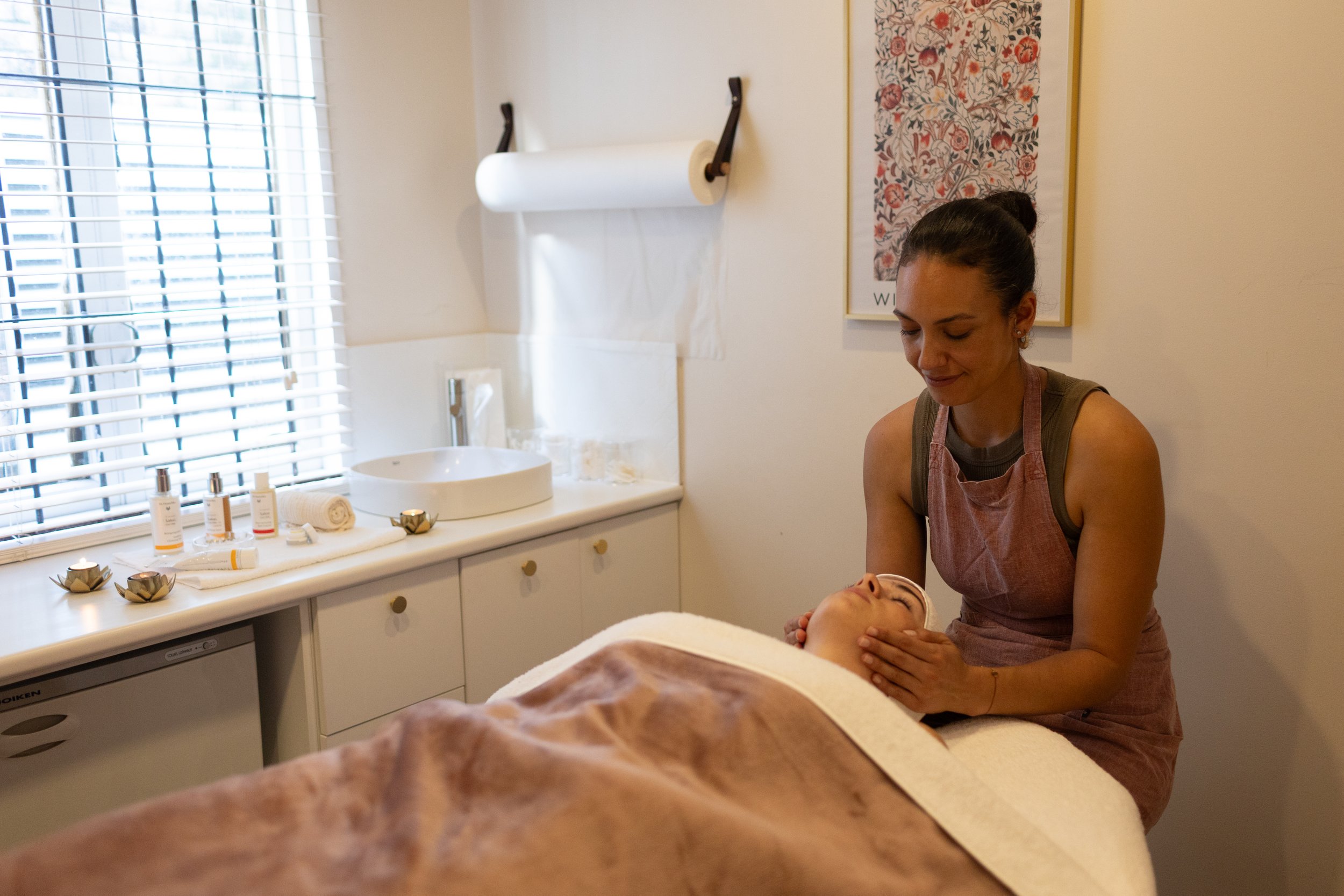 A woman giving a facial treatment to a client lying on a massage table in a spa room.