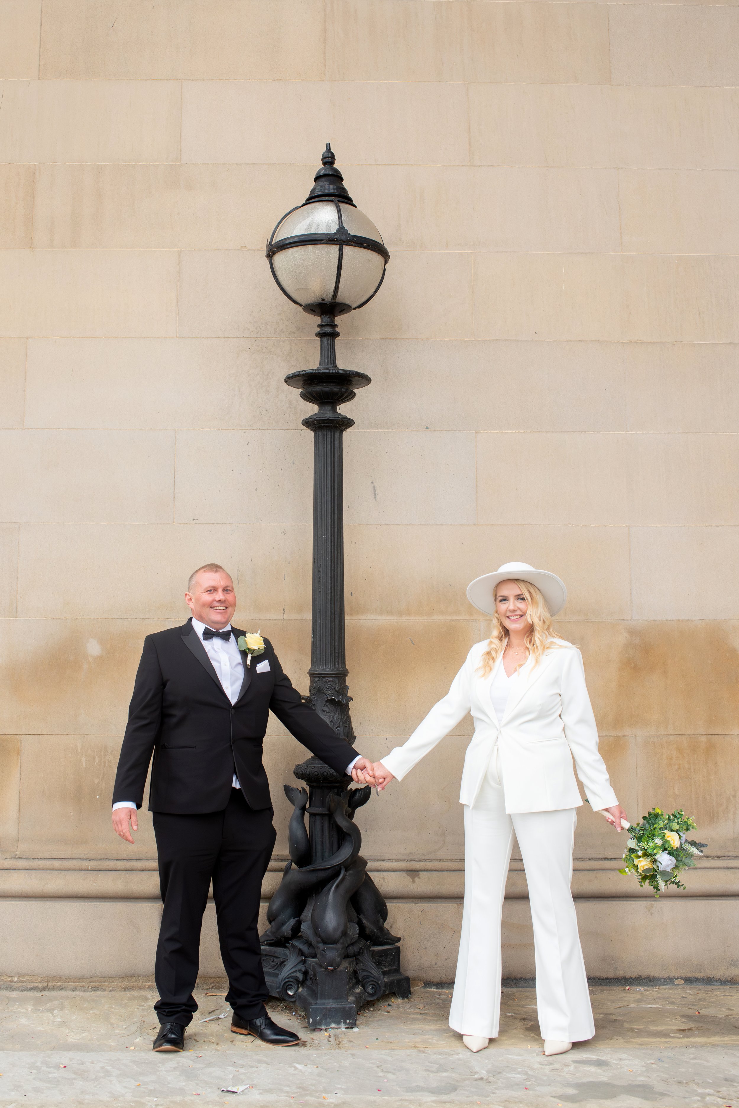 A bride and groom in wedding attire holding hands in front of a large streetlamp and a beige stone wall outside.