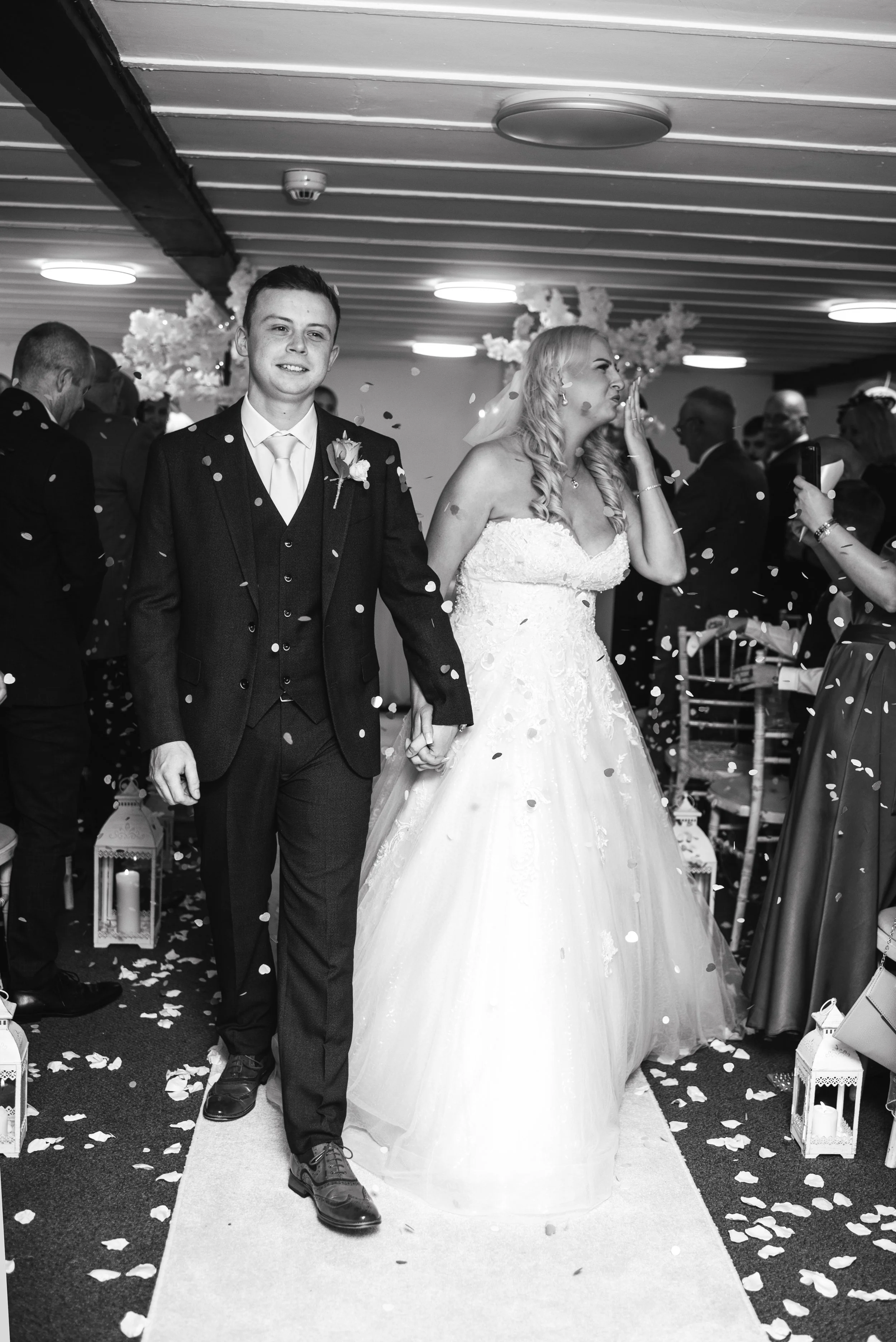 A bride and groom walking hand in hand down the aisle at their wedding ceremony, surrounded by wedding guests, with confetti falling around them.