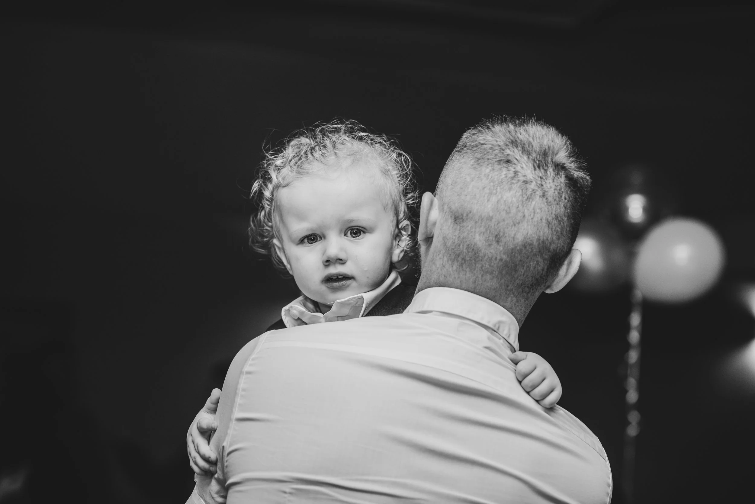 A little girl with curly hair looks directly at the camera while being carried by a man with short hair, seen from the back, against a dark background with balloons.