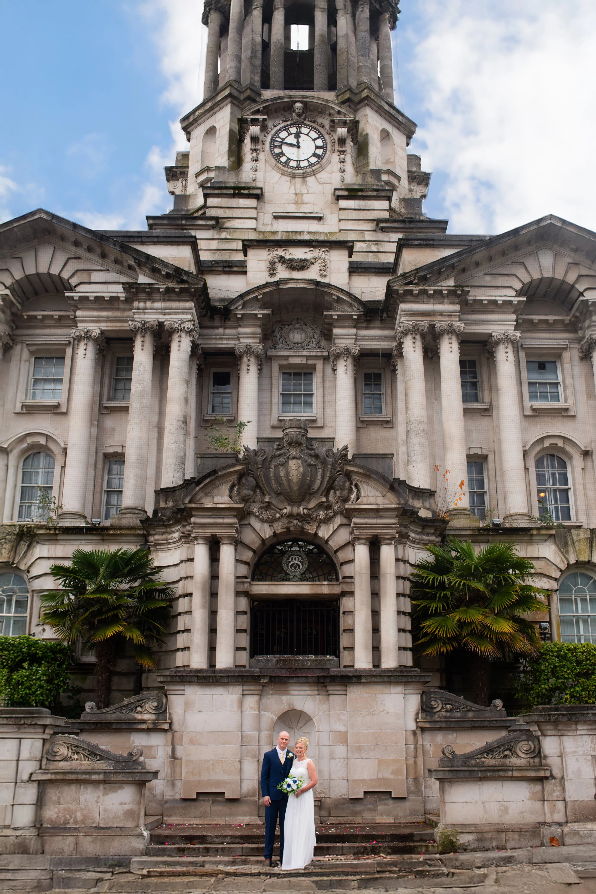 A wedding couple standing in front of a historic stone building with columns, decorative carvings, and a clock tower under a partly cloudy sky.