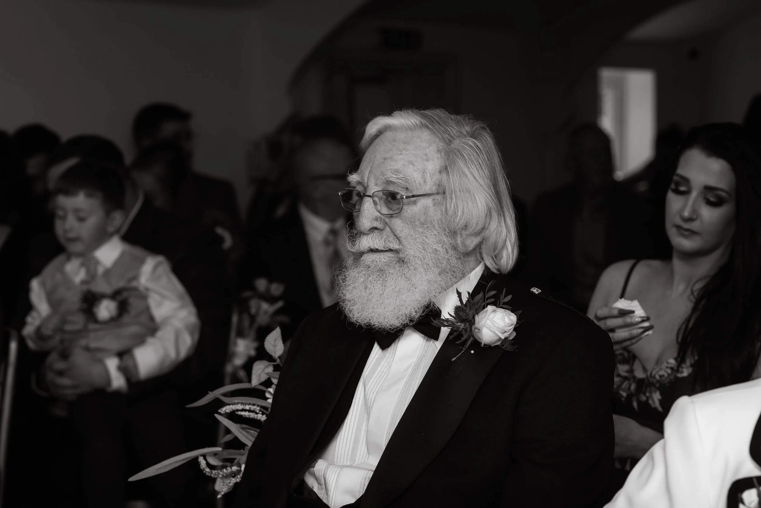 Older man with long white hair, beard, and glasses wearing a tuxedo with a boutonniere, sitting at a formal event, surrounded by other attendees including a young boy and a woman, in black and white.