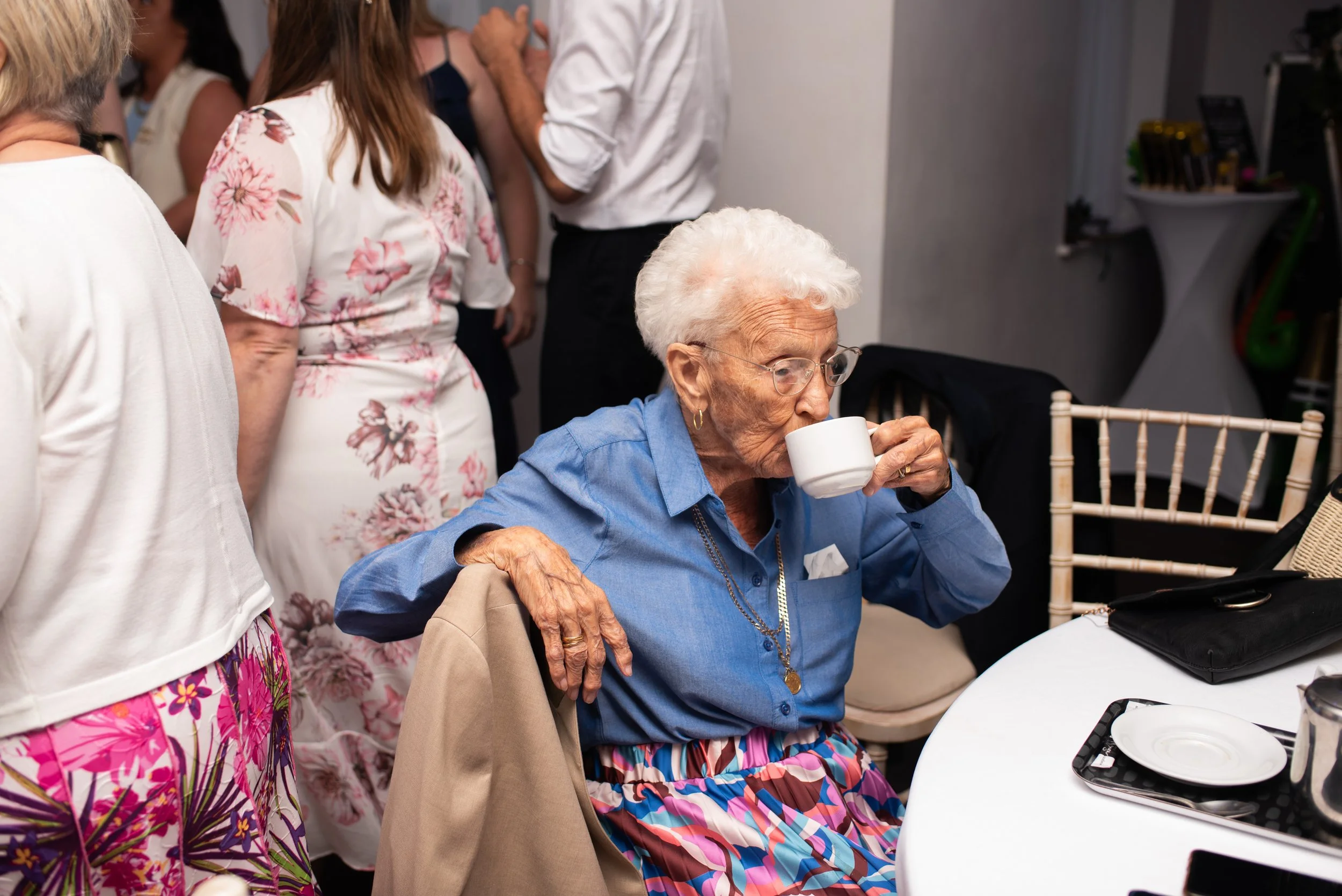 An elderly woman with white hair, glasses, and a blue shirt is sitting at a table, drinking from a white mug. Other people are standing nearby, engaged in conversation, at what appears to be a social gathering or party.
