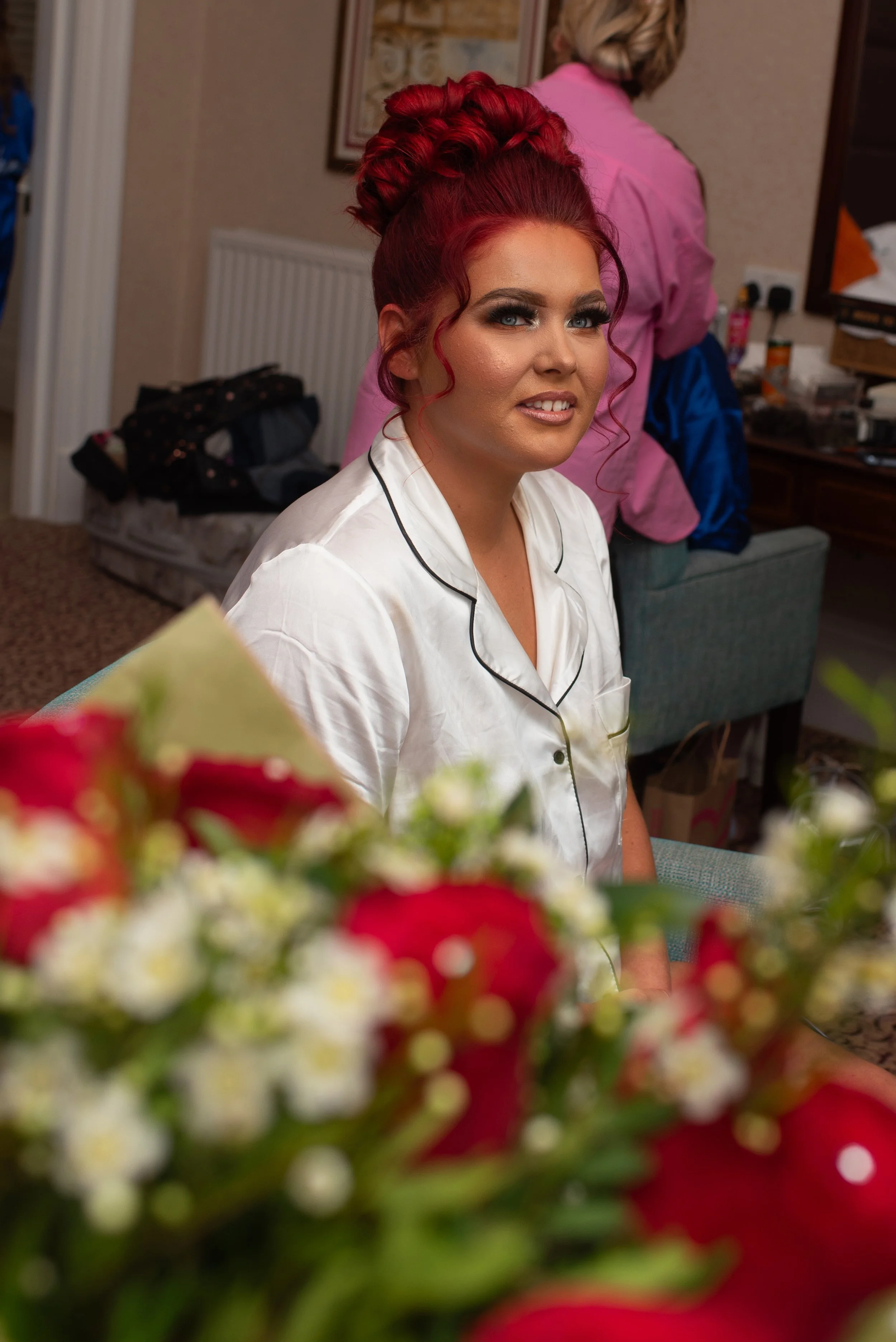 A woman with red hair styled in an intricate updo, wearing white pajamas with black piping, sitting in a room with a floral bouquet in the foreground and another person in a pink shirt in the background.