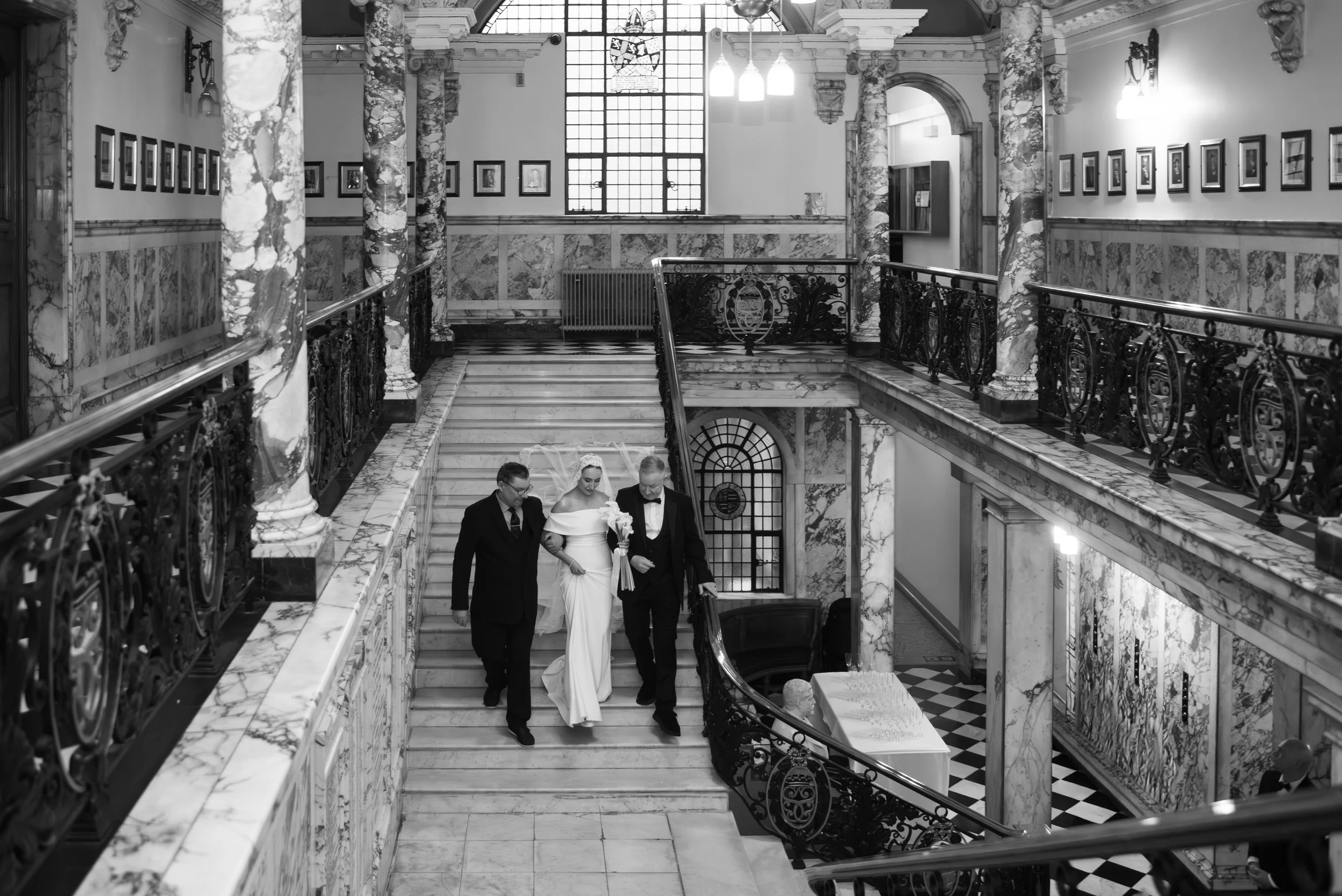 A bride in a white wedding gown holding a bouquet, accompanied by two men in suits, walking down marble stairs inside a grand, ornate building with marble columns and decorative railings.